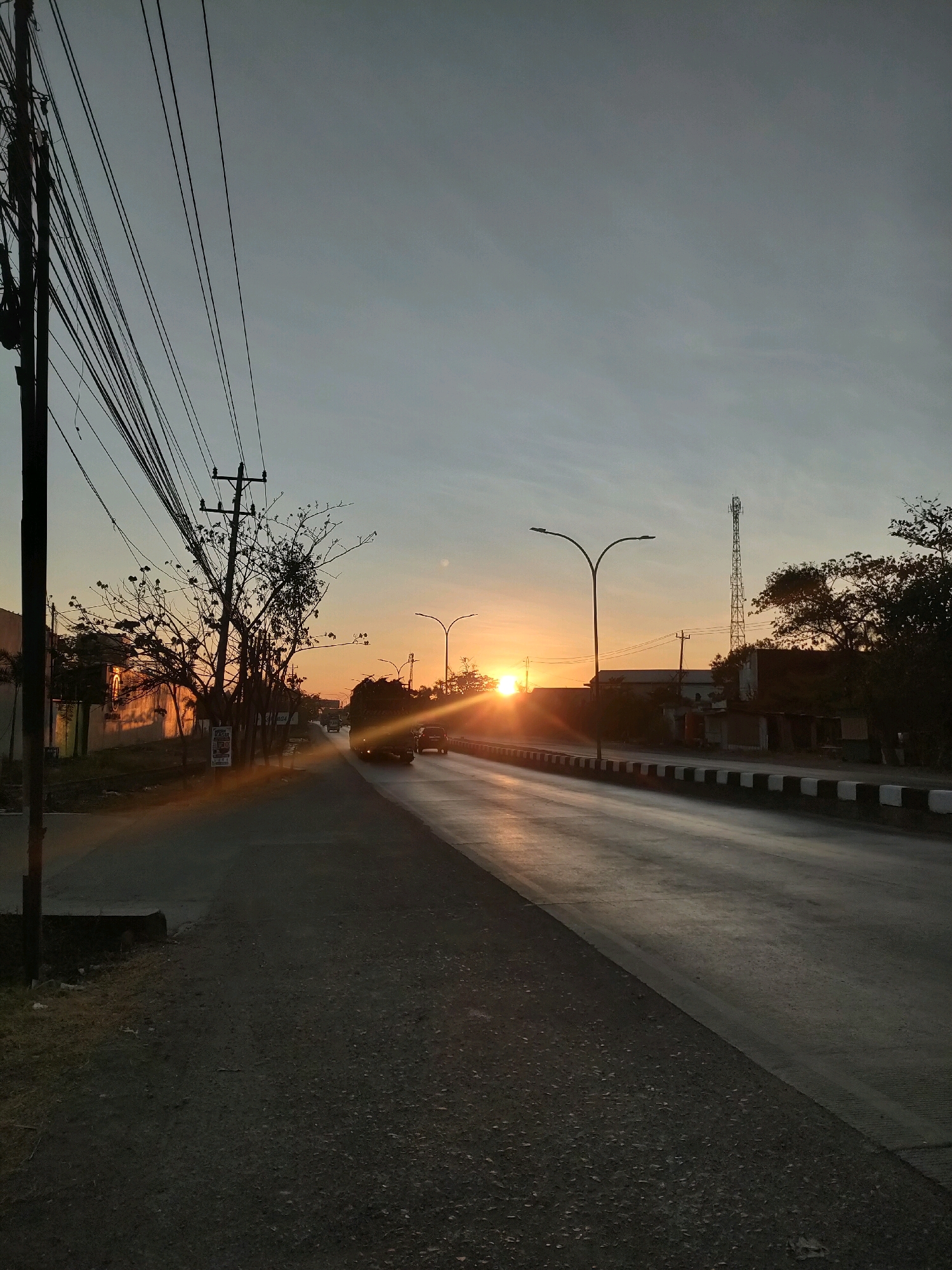 The image captures a street scene in Semarang, Indonesia, during what appears to be a sunrise or sunset. A road stretches into the distance, with vehicles visible. The sky is a muted blue-gray, with the sun a bright orange disc near the horizon, casting a warm glow and long shadows across the asphalt. On the left side of the road, there are power lines strung between poles, a bare-branched tree, and the side of a building. On the right side of the road, there are streetlights, a cell tower, and more trees. The road is lined with a raised curb featuring black and white markings. The overall atmosphere suggests an urban environment at the transitional time of day.