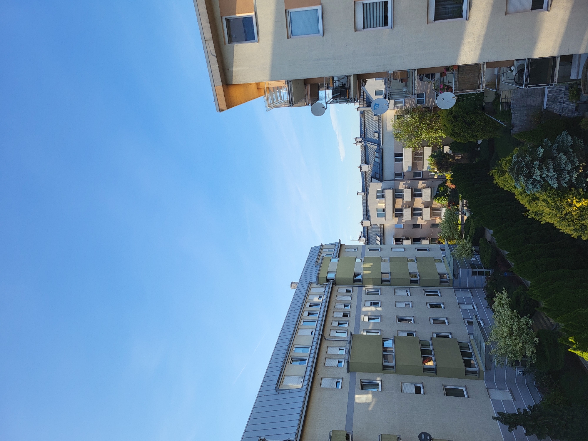 The image shows a low-angle view of several apartment buildings in an urban environment. The sky is clear and blue. The buildings are a mix of architectural styles and colors. Some have satellite dishes on their balconies. There is also a green area with trees and shrubs between the buildings. A grey walkway is visible alongside a multi-story building. The location is unknown.