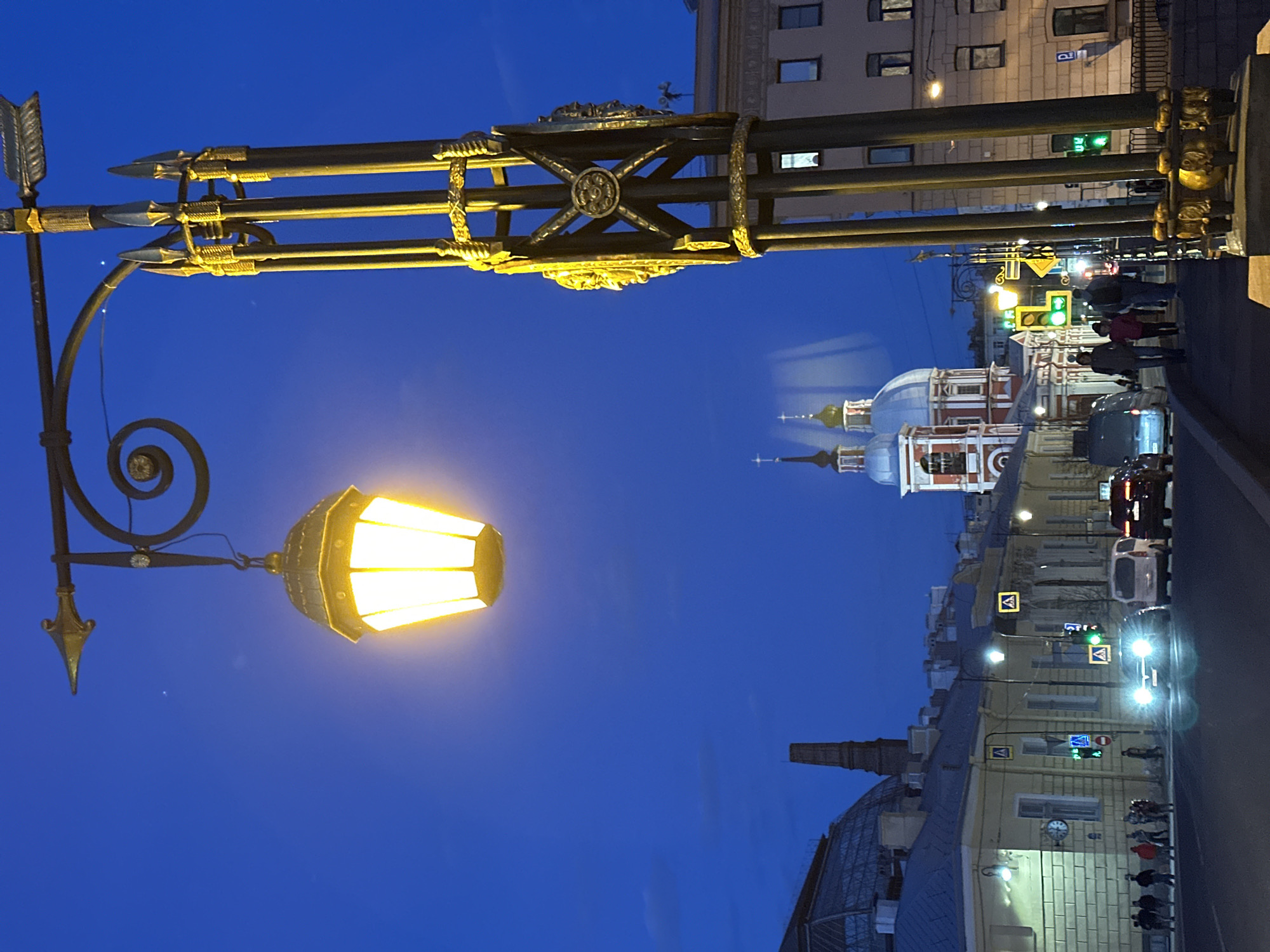 The image depicts a street scene in Saint Petersburg, Russia, at dusk or night. A vintage-style lamppost with a bright, glowing lantern is prominently featured in the foreground. Behind the lamppost, there is a view along a street. Buildings line the street, including one with a distinctive red and white dome. Pedestrians are walking along the sidewalk, and cars are driving on the street. Streetlights and traffic signals are visible, indicating an urban setting. The sky is a deep blue color. The architectural details suggest a historical or culturally significant area within the city.