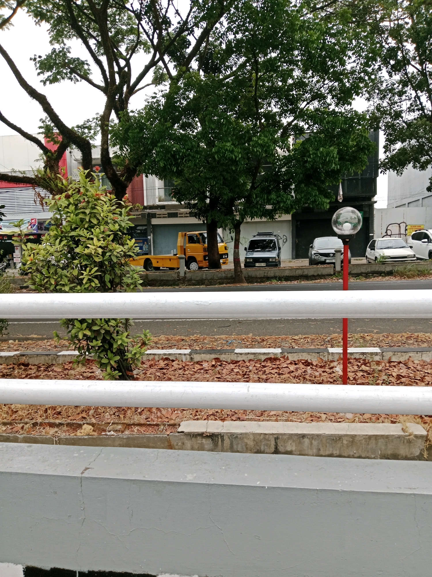 The image depicts a street scene in Muka, Indonesia, focusing on a roadside environment with a focus on a white railing in the foreground. Behind the railing, a street is visible with several vehicles parked and driving. These include a yellow flatbed truck, a dark-colored van, a silver car, and a white car. The street is lined with trees, one with a large, sprawling canopy, and some smaller bushes. A street lamp with a spherical glass shade is also present, mounted on a red pole. The ground beside the street is covered with fallen brown leaves and some dry grass. Buildings are visible in the background, appearing to be commercial establishments, with signage that is partially obscured. The overall atmosphere suggests an urban or suburban setting with natural elements integrated into the built environment.