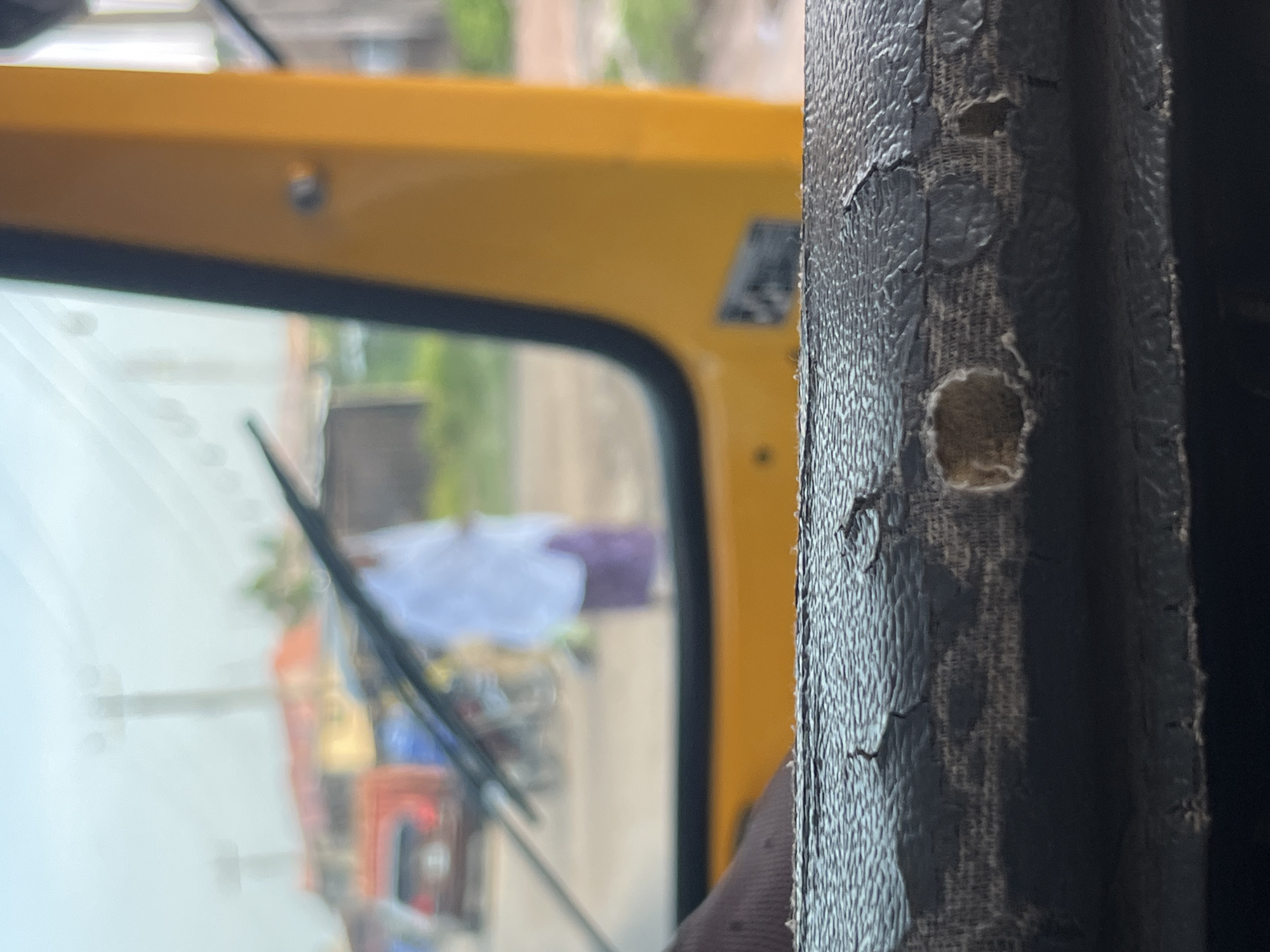 The image is taken from inside a yellow vehicle, likely a tuk-tuk, looking out through the windshield. In the foreground, the worn and cracked dashboard of the vehicle is visible, with a dark fabric covering. Through the windshield, a street scene is depicted, blurred due to the distance and focus. A person wearing a white top and a purple skirt or lower garment stands in the middle ground. Behind them, various objects, possibly vehicles and buildings, are present but indistinct. The environment appears to be an urban or semi-urban setting with some greenery and what seem to be utility poles. The sky is overcast.