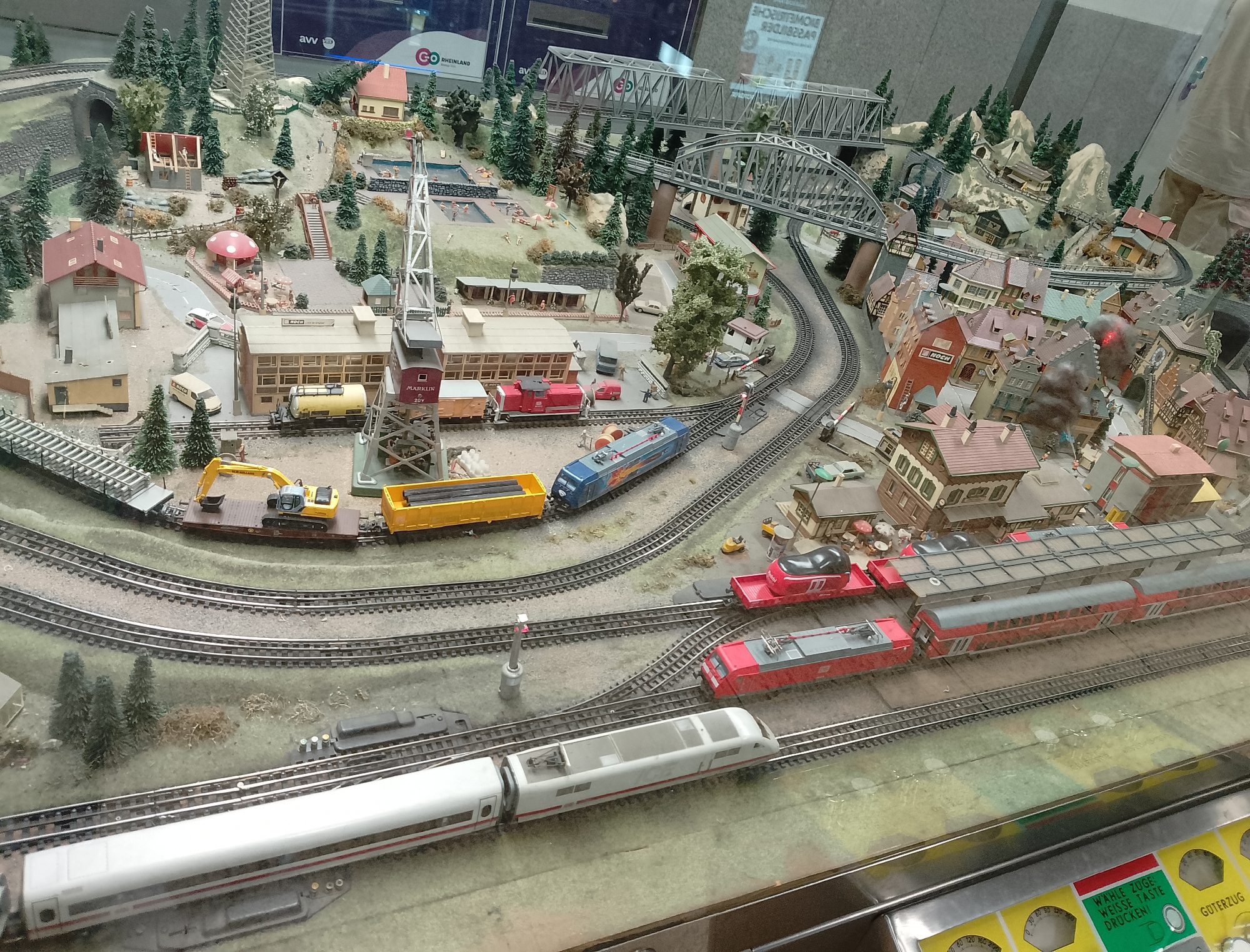 This is a high-angle, medium shot of a detailed model train diorama. The diorama depicts a miniature town with buildings, trees, train tracks, and various vehicles. In the foreground, a long, white train with red accents runs on a track. Behind it, a red and gray electric locomotive is positioned on another track, with a red freight car attached. Further back, a blue train is seen on a curved track that goes under a bridge. The diorama includes a variety of structures, including houses with red roofs, a church-like building with a tall spire, and a crane. There are also elements of the natural landscape, such as miniature trees and hills. Some areas of the diorama appear to be designed to represent water, such as a swimming pool with small figures. The scene appears to be set during the daytime, with no indication of weather conditions. Visible text on signage includes "MARKLIN," "NOCH," "CO RHEINLAND," and German phrases like "WAHLE ZUGE WEISSE TASTE DRUCKENI" and "GUTERZUG." The overall impression is a meticulously crafted and complex model railway.