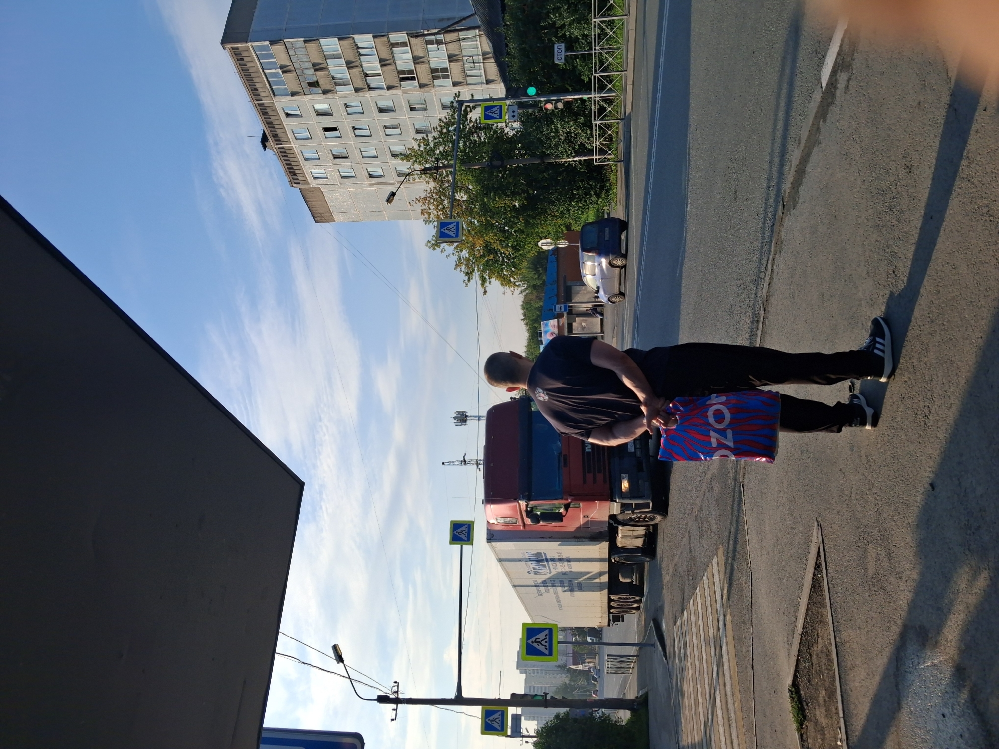 A man stands on a sidewalk holding a red, blue, and white patterned shopping bag. He is wearing a black t-shirt and black pants with white stripes on the sides, and black and white athletic shoes. He is facing away from the camera, looking at a red truck driving on a road. The truck is hauling a white trailer. There are several pedestrian crossing signs visible. In the background, there is a tall, light-colored apartment building. The sky is blue with some clouds, and sunlight is casting shadows on the ground. A blue car is parked on the side of the road. The scene appears to be a street in a city. The shopping bag has the word "OZON" printed on it.