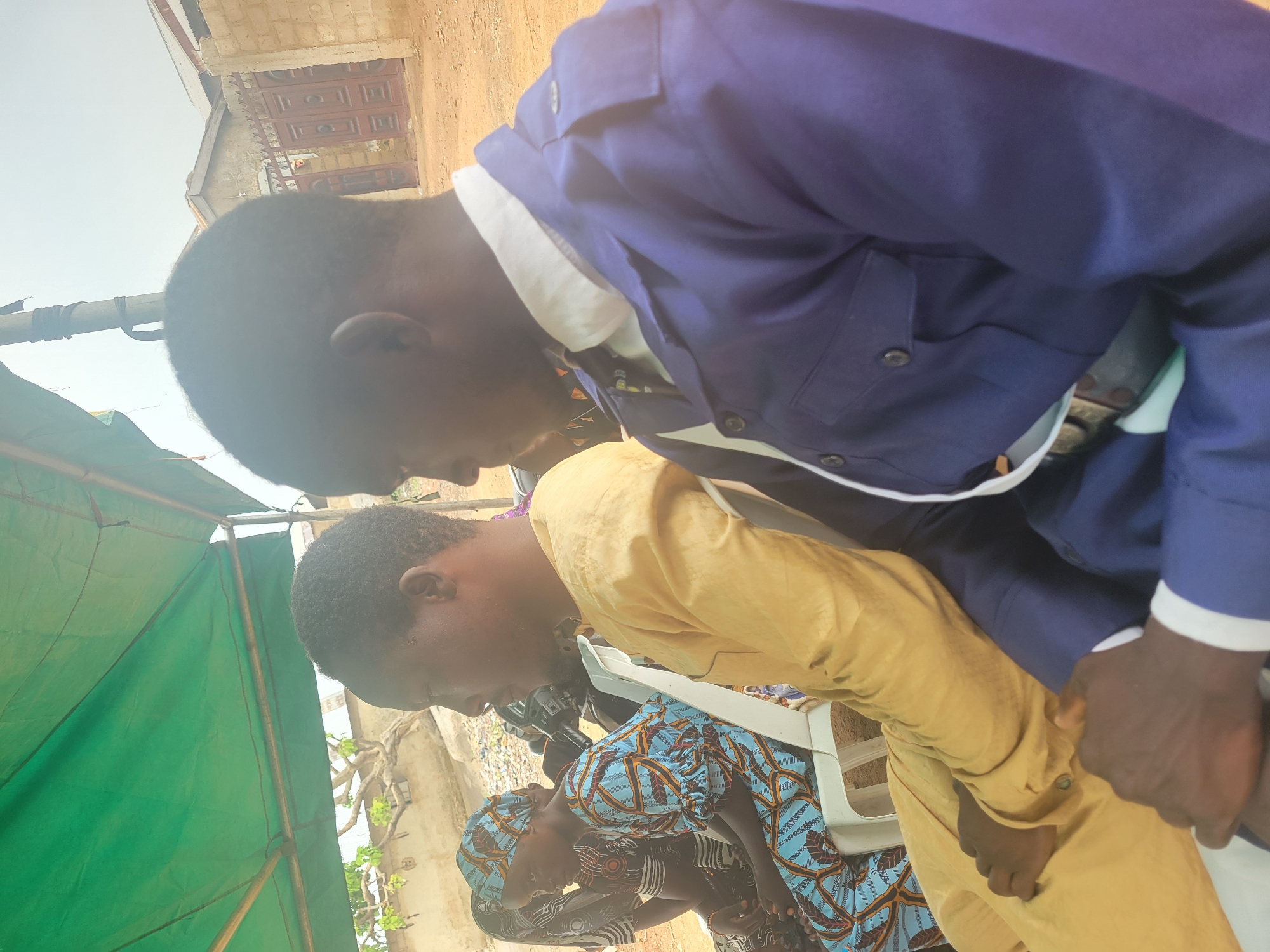 In Madalla, Nigeria, the image captures a candid moment outdoors. Two men are prominently featured: one in a blue suit and the other in a gold tunic. Both have their heads bowed in the same direction. A woman wearing a patterned blue and brown outfit is seated nearby, also with her head lowered. The scene appears to be set under a makeshift green tent. In the background, the architecture of a building is visible.