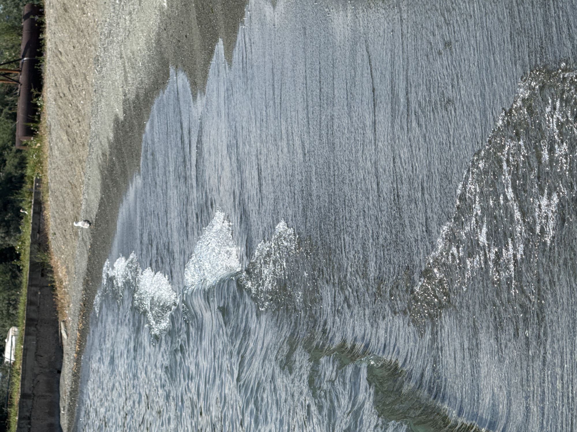The image depicts a shoreline scene in Tsamakaberd, Armenia, under natural daylight. The foreground is dominated by the surface of a body of water, likely a lake, with gentle waves lapping against a pebbled shore. The water is a muted blue-grey, with the sunlight creating shimmering highlights on the crests of the small waves. The shoreline itself is composed of small, dark grey pebbles mixed with sand, and slopes upwards towards a grassy embankment. In the background, a concrete wall runs along the edge of the embankment, with some sparse vegetation and trees growing behind it. Further in the distance, a large, rusty cylindrical object, possibly industrial equipment, is visible amongst the greenery. A single white bird, likely a seagull, stands on the pebbled shore. The overall impression is one of a natural, somewhat rugged coastal environment.