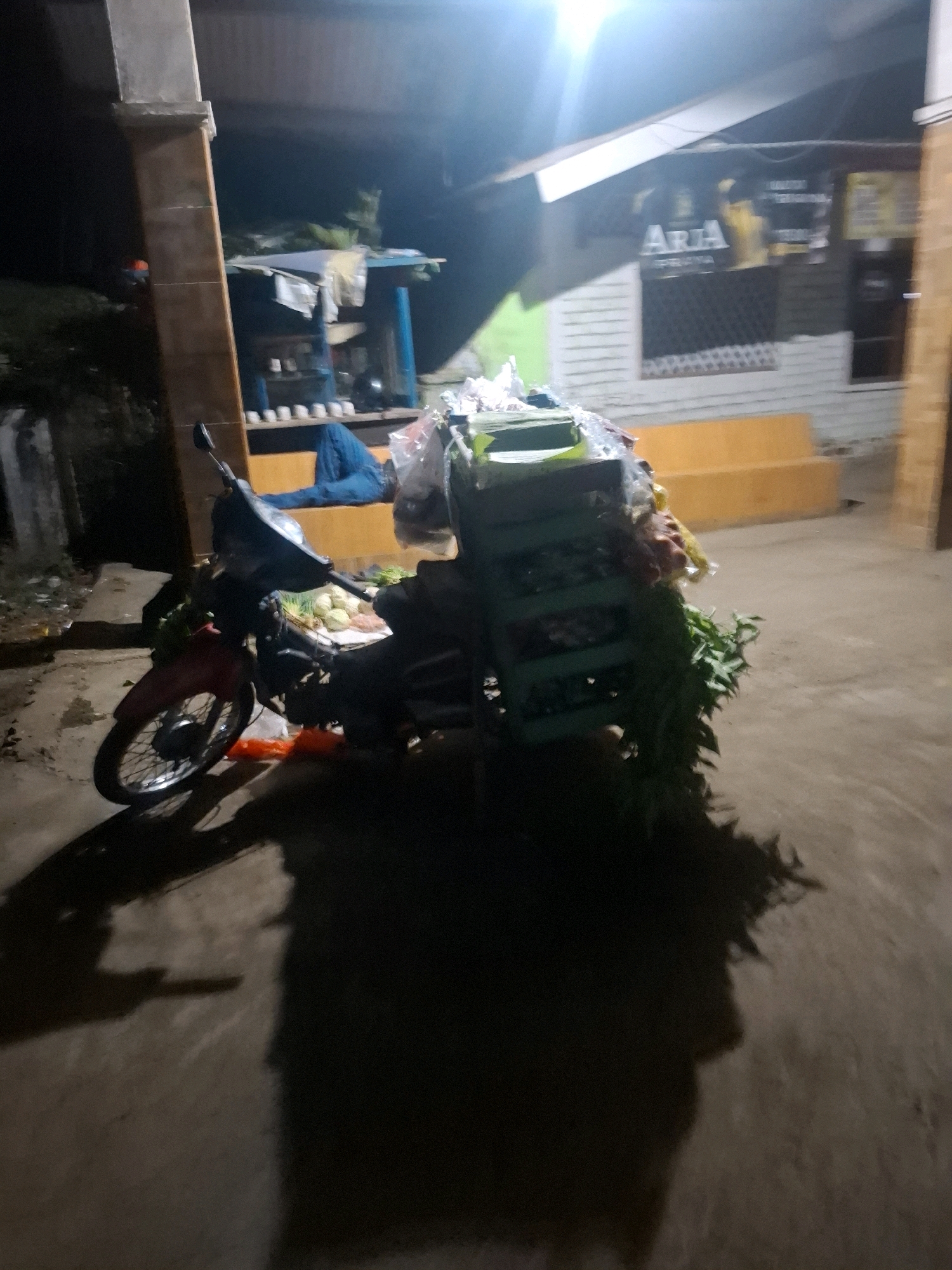 The image depicts a nighttime scene on a street in Ciheulang Tonggoh, Indonesia. A motorcycle is parked, loaded with various goods, including what appear to be vegetables and other produce, contained in crates and plastic bags. The motorcycle is situated in front of a building with a sign that reads "ARIA." To the left of the motorcycle, there is a structure with pillars, and to the right, an orange-colored bench is visible. The ground is paved, and the lighting suggests it is dark, with artificial light illuminating the scene. Shadows of the motorcycle and possibly people are cast on the ground. A portion of a blue tarp is also visible near the building.