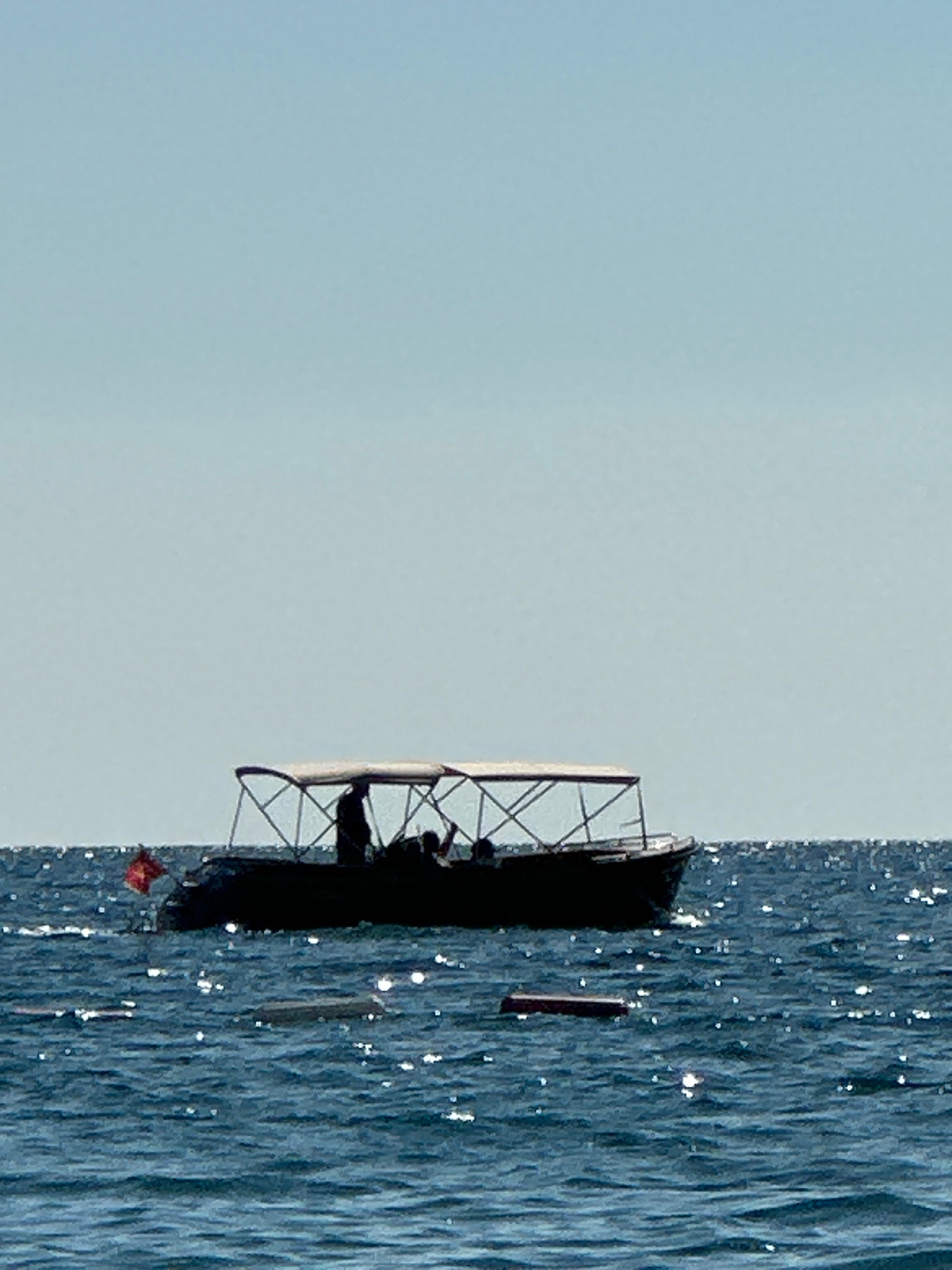 In the city of Boreti, Montenegro, a dark-hulled boat with a white canopy is visible on the blue, rippling sea under a clear sky. The boat is populated by silhouetted figures. A small red flag is attached to the stern of the boat. Several rectangular objects are floating on the water's surface in front of the boat, and the sun creates bright reflections on the water.