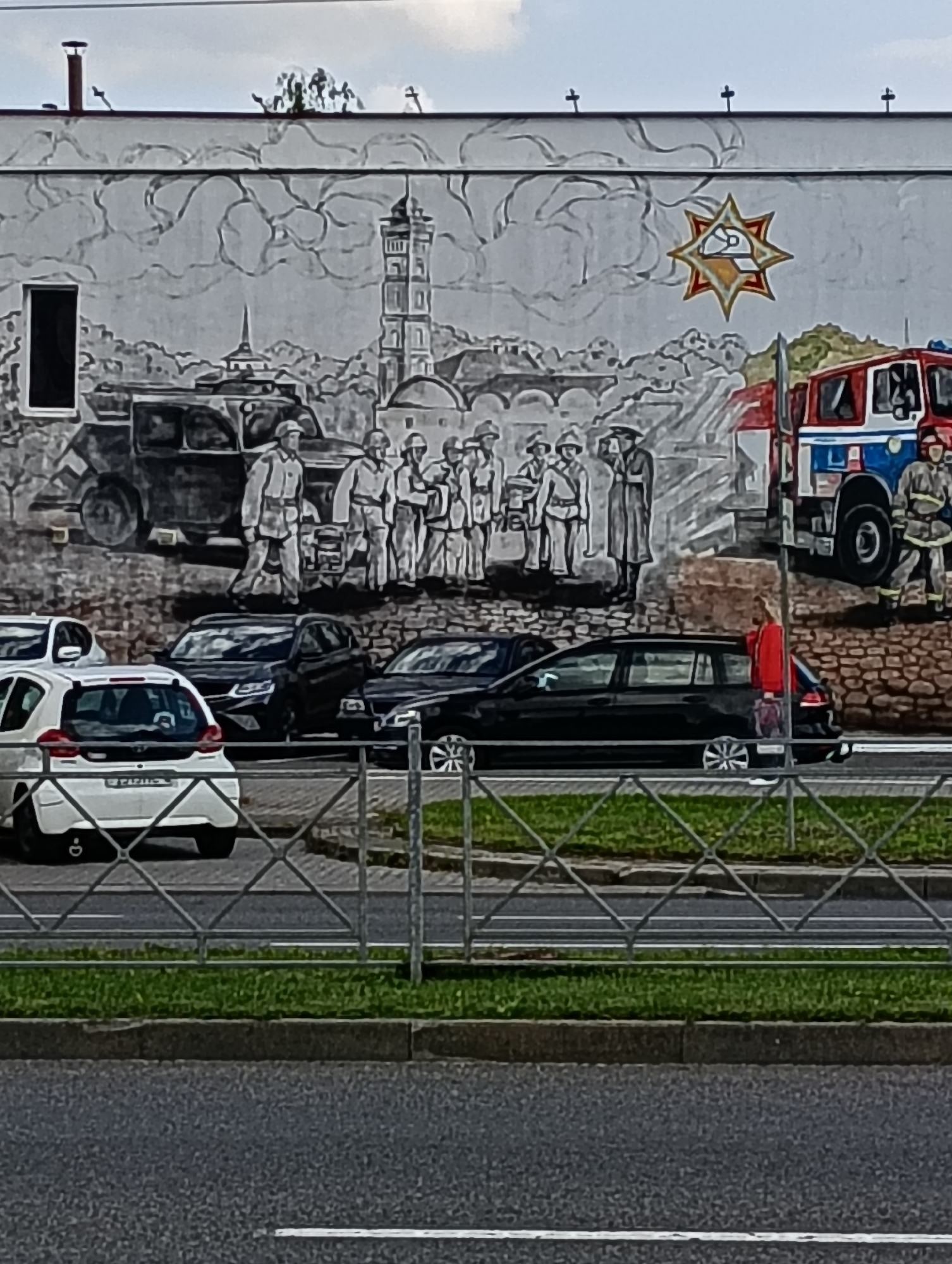The image captures a mural on the side of a building in Hrodna, Belarus, depicting a historical scene with firefighters. In the foreground, several cars are parked on a street, with a metal fence separating the street from a grassy area and the building. The mural itself is rendered in grayscale and features a fire truck, several figures in uniform, and a prominent star emblem. The building has a few visible architectural details, including a window and roofline elements. The sky above is overcast.