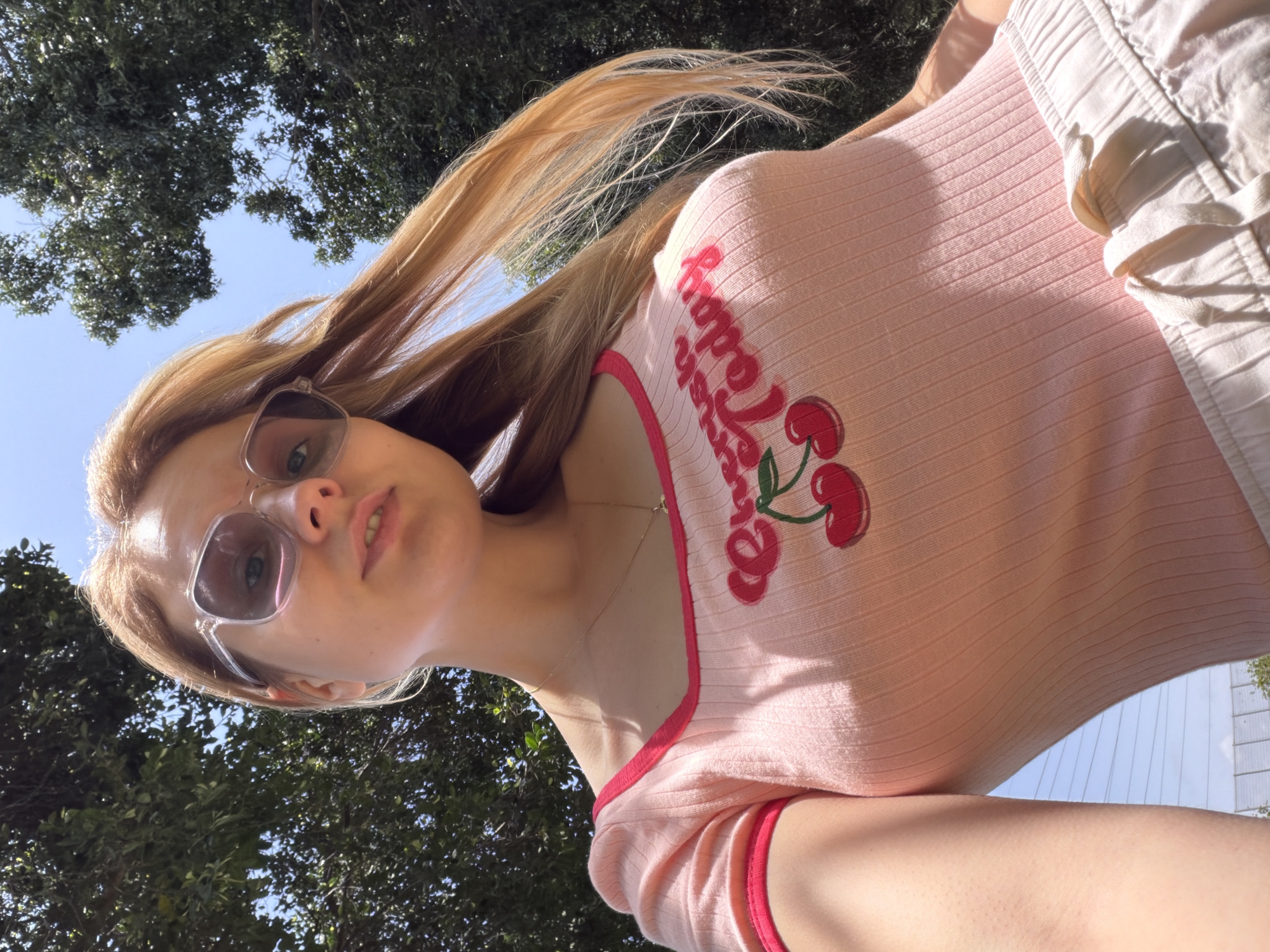 A young woman is pictured from a low angle, looking directly into the camera. She is wearing sunglasses with transparent frames and a light pink, ribbed t-shirt with a "Happy Today" and cherry motif. She is wearing light-colored shorts. Her blonde hair is swept to the side. The background is a mix of green foliage, suggesting trees, and a glimpse of a bright blue sky. It's difficult to confirm if the location is in Tel Aviv, Israel, based solely on the visual information provided.