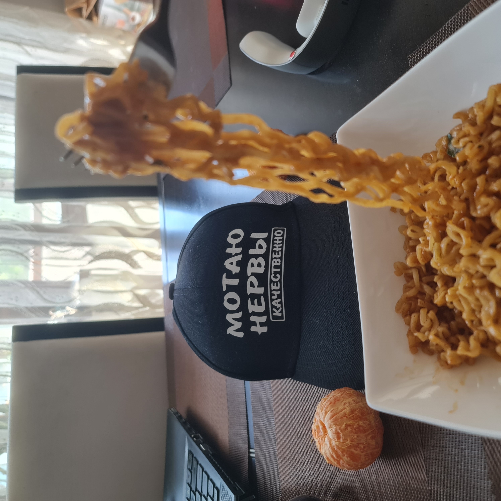The scene takes place indoors, in the city of Kemerovo, Russia, captured from a slightly elevated, close-up perspective, focusing on a meal. In the foreground, a white rectangular bowl is filled with cooked, sauced instant noodles. A metal fork, held by an unseen individual, lifts a substantial portion of these noodles out of the bowl and extends them towards the top right of the frame, indicating the act of eating.
The table surface is dark and reflective, partially covered by a brown, woven placemat beneath the bowl. To the left of the bowl, resting on the placemat, is a single, peeled orange tangerine. Further to the left, partially visible, is the keyboard and base of an open black laptop.
Centrally positioned on the table, behind the bowl and tangerine, is a black baseball cap. The front of the cap features white Cyrillic text: "МОТАЮ НЕРВЫ" (Motayu Nervy), which translates to "I get on nerves," and below it, "КАЧЕСТВЕННО" (Kachestvenno) in a framed box, translating to "With quality" or "Expertly." The combined message is "I expertly get on nerves."
In the mid-ground, a white and black electronic device with a red accent is visible on the dark table surface to the right of the noodles. The background features two light-colored (beige or off-white) dining chairs with dark vertical accents, positioned in front of a window. Sheer, patterned curtains are drawn across the window, allowing bright, natural daylight to illuminate the scene and providing a soft, diffused view of exterior greenery.