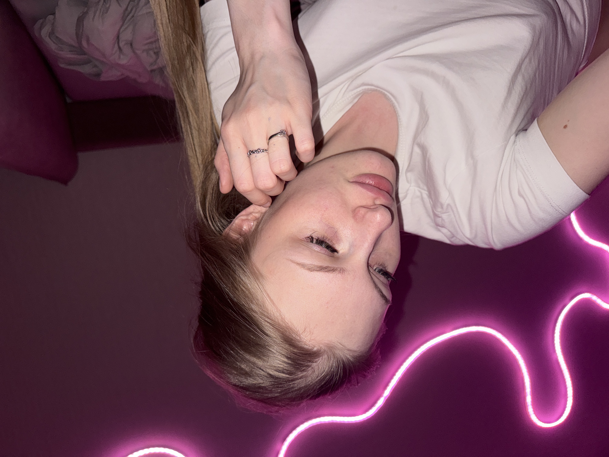 The image depicts a close-up, indoor shot of a young woman with light brown hair, wearing a white t-shirt. She is positioned in the foreground, with her head turned slightly to the left, exposing her profile. Her right hand is raised to her face, with two silver rings visible on her fingers. A pink neon light strip is present in the background, forming a curved shape against a dark purple wall. The overall ambiance suggests a dimly lit, possibly bedroom setting. The image does not provide any specific identifiable landmarks or context that would allow for pinpointing a location within the city of Perm, Russia.