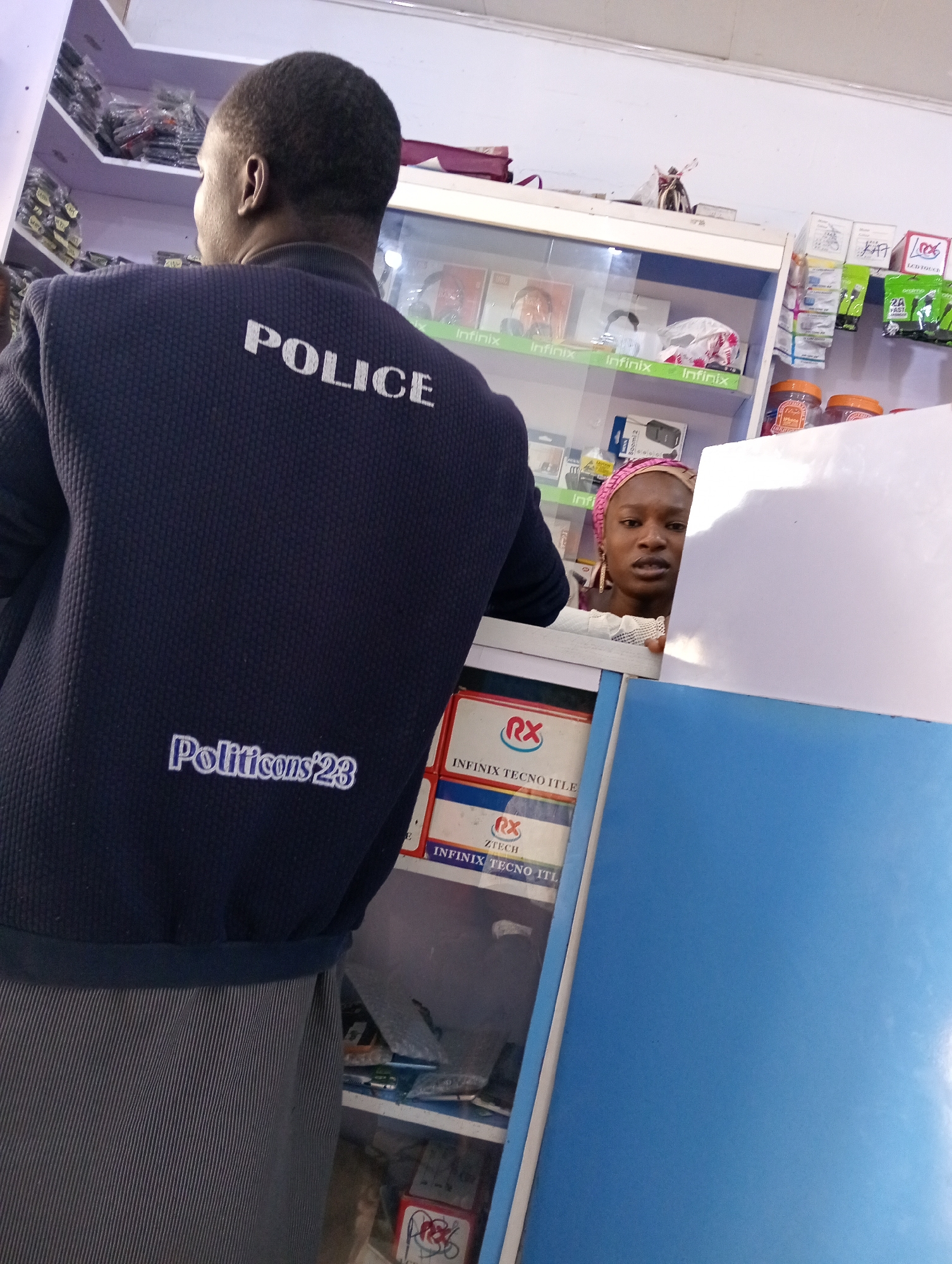 A photograph taken indoors shows a police officer in uniform with "POLICE" written in white block letters across the upper back, along with "Politicons 23" in script font just above the waistline of his dark blue jacket. The officer's back is to the camera, and he stands near a counter in what appears to be a retail shop. On the right side of the frame, a woman with a pink headscarf looks out from behind the counter, near stacked product boxes for "INFINIX TECNO ITLE" and "ZTECH." Shelves in the background are stocked with various electronics and packaged goods. The location and time of the photo are unknown.