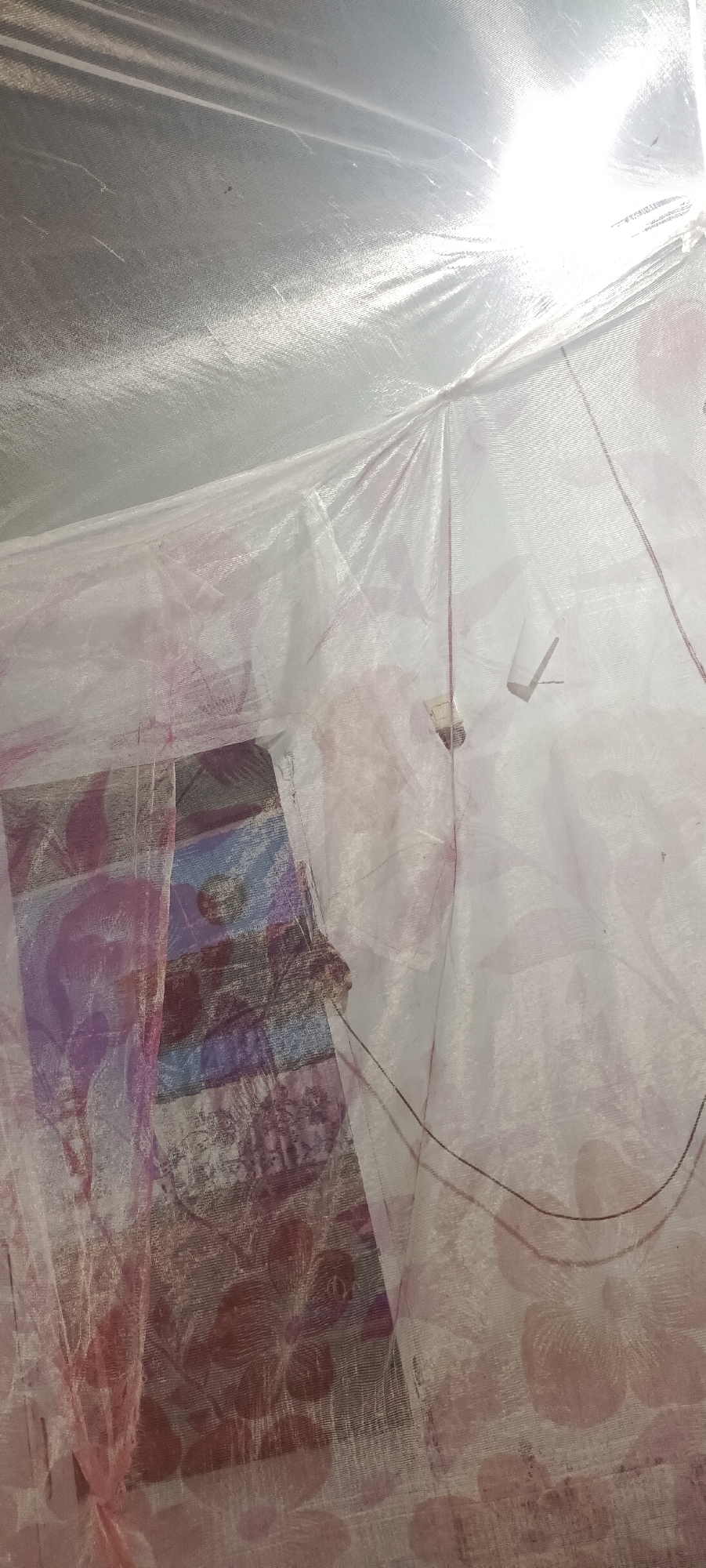 This image shows a close-up view of a mosquito net with a floral pattern, possibly in a bedroom or living space. The net is white and translucent, with subtle pink floral motifs. A bright light source, likely a ceiling light or a lamp, is visible at the top of the image, causing significant glare and washing out details in that area. There is a rectangular object with horizontal stripes of color, appearing to be some sort of fabric or tapestry, visible through the net on the left side of the image. Thin black wires are also visible, possibly from a light fixture or a fan, trailing across the net. The overall impression is of a dimly lit, domestic interior. There is no indication of the city of Damuli Pekan, Indonesia.