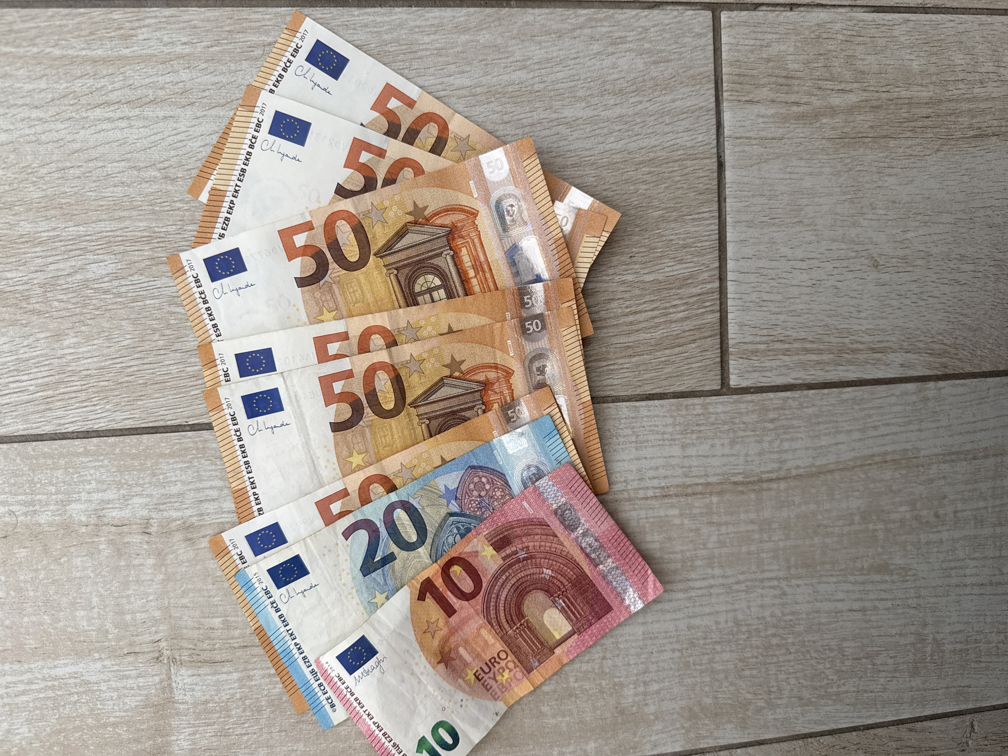 The image shows a collection of Euro banknotes spread out on a light-colored, wood-patterned tiled surface. The banknotes are fanned out, with the denominations visible being 10, 20, and 50 Euros. There are at least three 50 Euro notes, one 20 Euro note, and one 10 Euro note. The 50 Euro notes appear to be newer issues, indicated by the year "2017" visible on them. The 10 Euro note is pink, the 20 Euro note is blue, and the 50 Euro notes are orange. The banknotes are laid out in a slightly overlapping manner. The surface they are on consists of rectangular tiles with a wood grain texture and visible grout lines between them. The lighting suggests an indoor setting. There is no indication of the city of Burgas, Bulgaria, in the image.