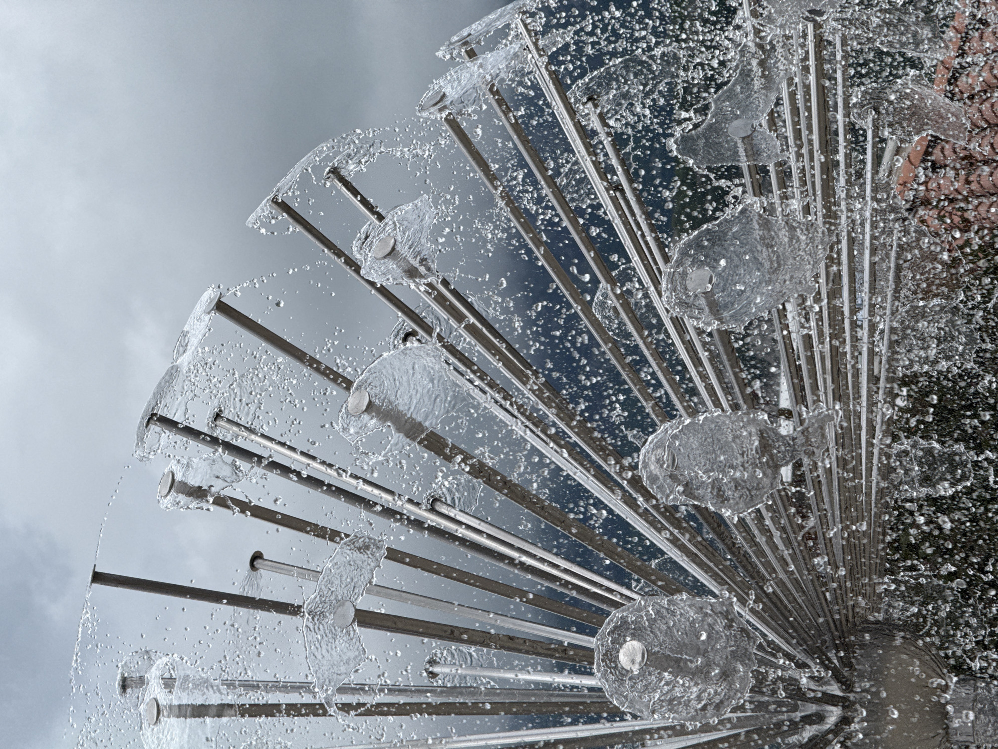 This image depicts a dandelion fountain located in Budva, Montenegro. Water is shown spraying from multiple nozzles on the metal structure. The background is a blurred mix of gray sky and foliage.