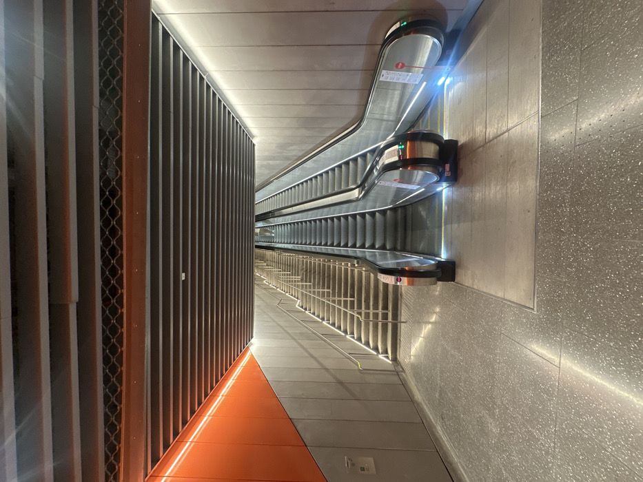 Three escalators ascend in parallel, brightly lit with integrated strips. Metal railings accompany both escalators and stairs. A metal wall with vertical slats and orange accents lines the left. The right side features a textured grey wall, possibly terrazzo. The lighting accentuates the sleek, modern design of the subway station. The space appears clean, sterile, and well-maintained. The scene evokes a sense of efficiency and movement.