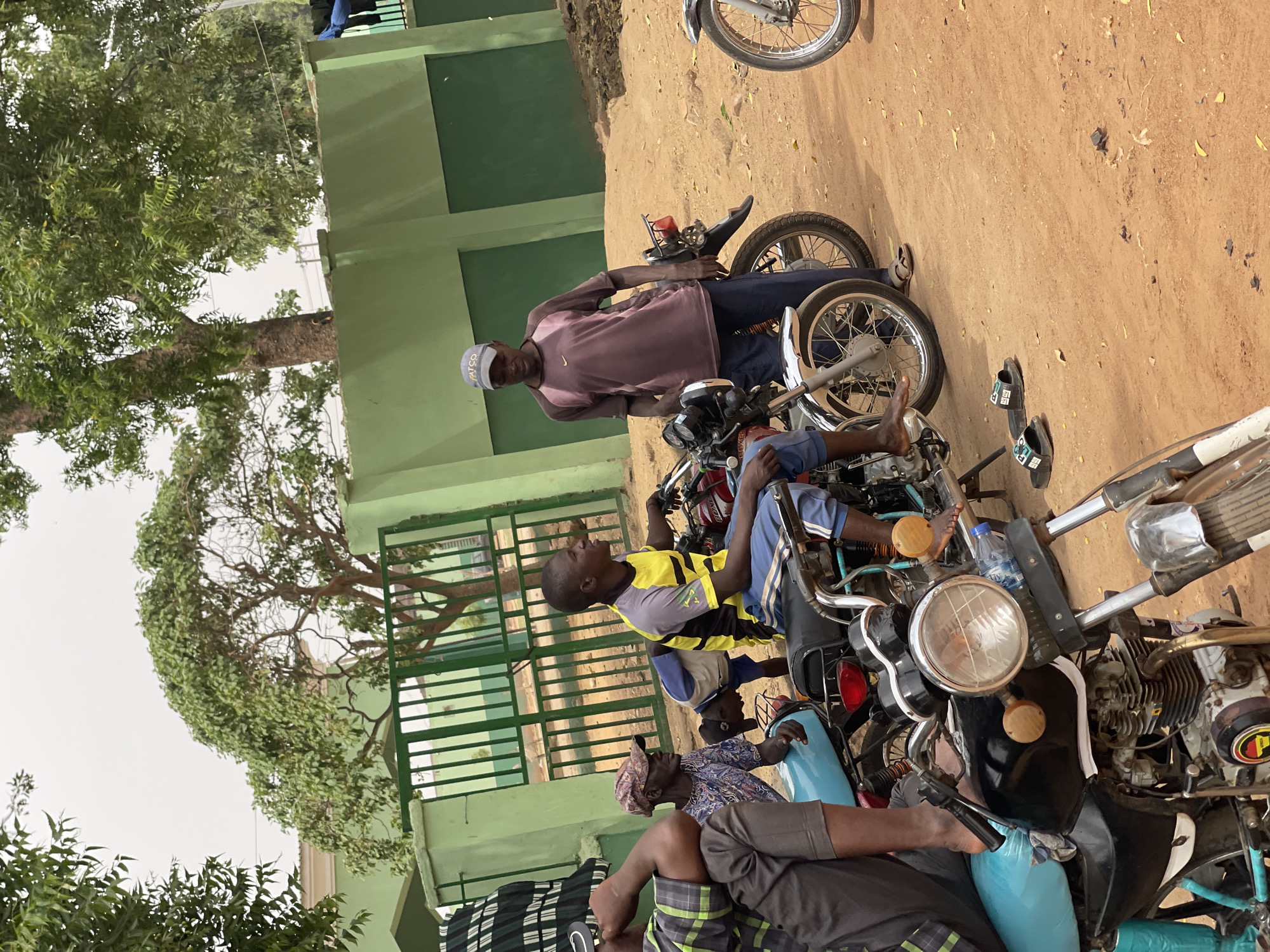 In Muchiya, Nigeria, a group of men gathers around several motorcycles on a dirt lot adjacent to a green building with a gated fence and a large tree. One man stands near a motorcycle, wearing a purple shirt and a light-colored cap. Another man is seated on a motorcycle, wearing a yellow and grey shirt. Additional people are seated on the other motorcycles. The ground is dirt, and there are leaves scattered across it.