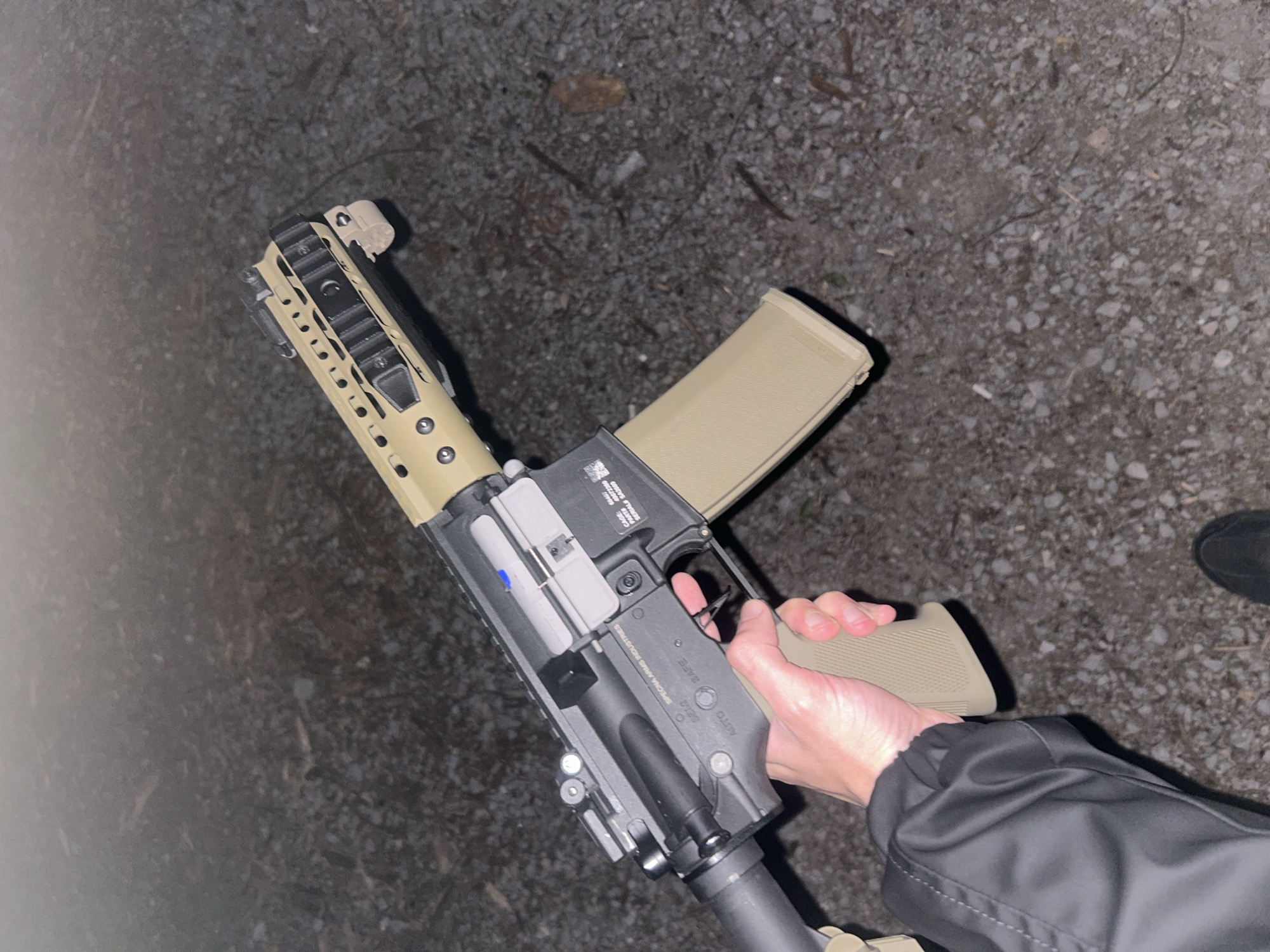 A person is holding an AR-15 style rifle outdoors on an unlit surface. The rifle is tan and grey, with a tan magazine. The person is wearing a black jacket. The surface appears to be gravel or small stones. No other people are visible. The location is not discernible as Veliko Tarnovo, Bulgaria.