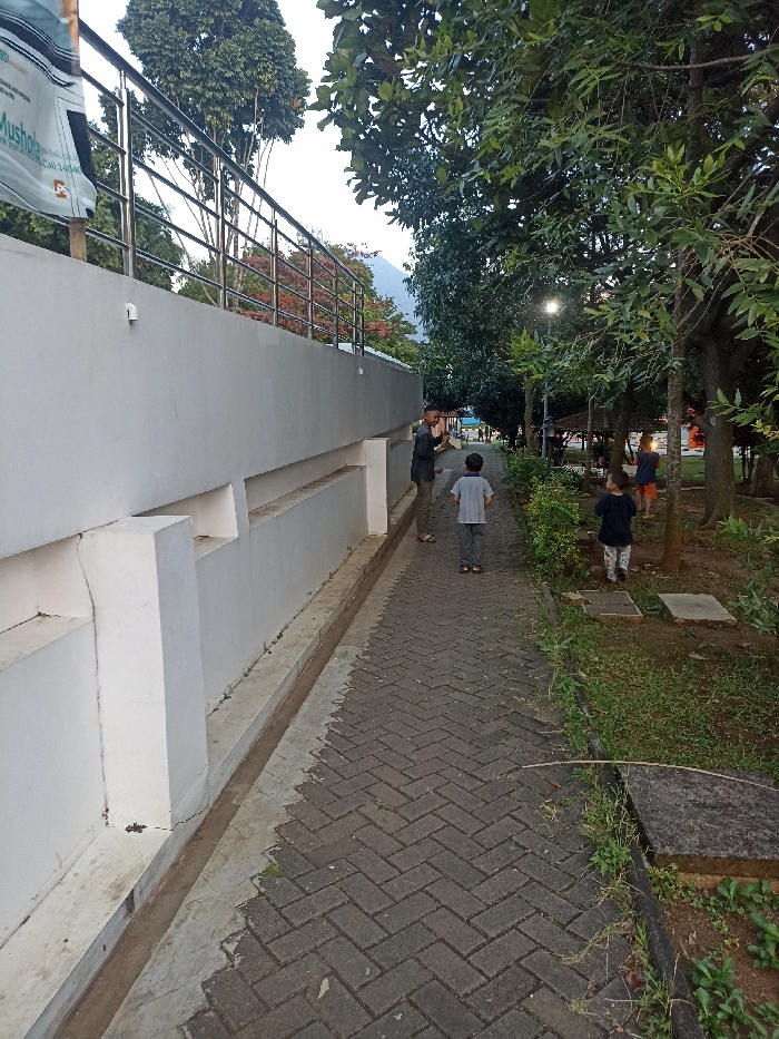 The image shows a paved walkway next to a white wall with a railing on top. There are three people walking on the walkway: two adults and one child. The child is walking behind the two adults. There is also a child standing on the grass next to the walkway. The environment is lush and green with plenty of trees and bushes. The location is Jatinangor, Indonesia, and the coordinates are -6.931 latitude and 107.776 longitude.