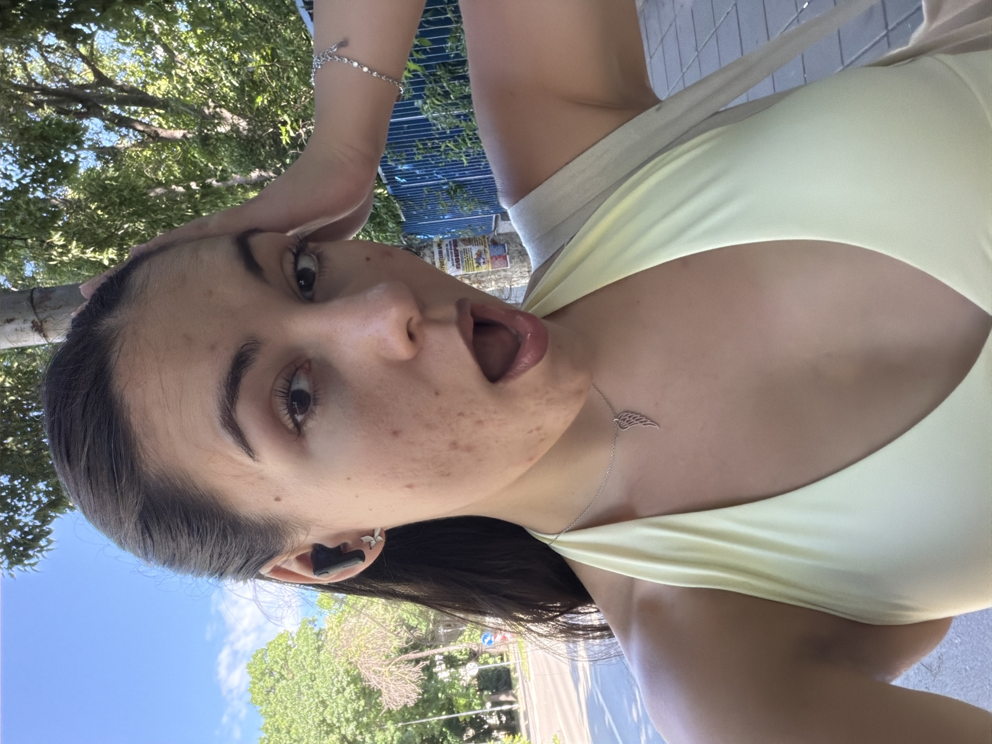 A young woman with dark hair is taking a selfie outdoors on a sunny day. She is wearing a light yellow halter top and a silver necklace with a wing pendant. She has an earbud in her left ear and is touching her forehead with her right hand. Her mouth is open in surprise or excitement. The background shows trees, a street, and a blue fence. The location is near a street in Varna, Bulgaria.
