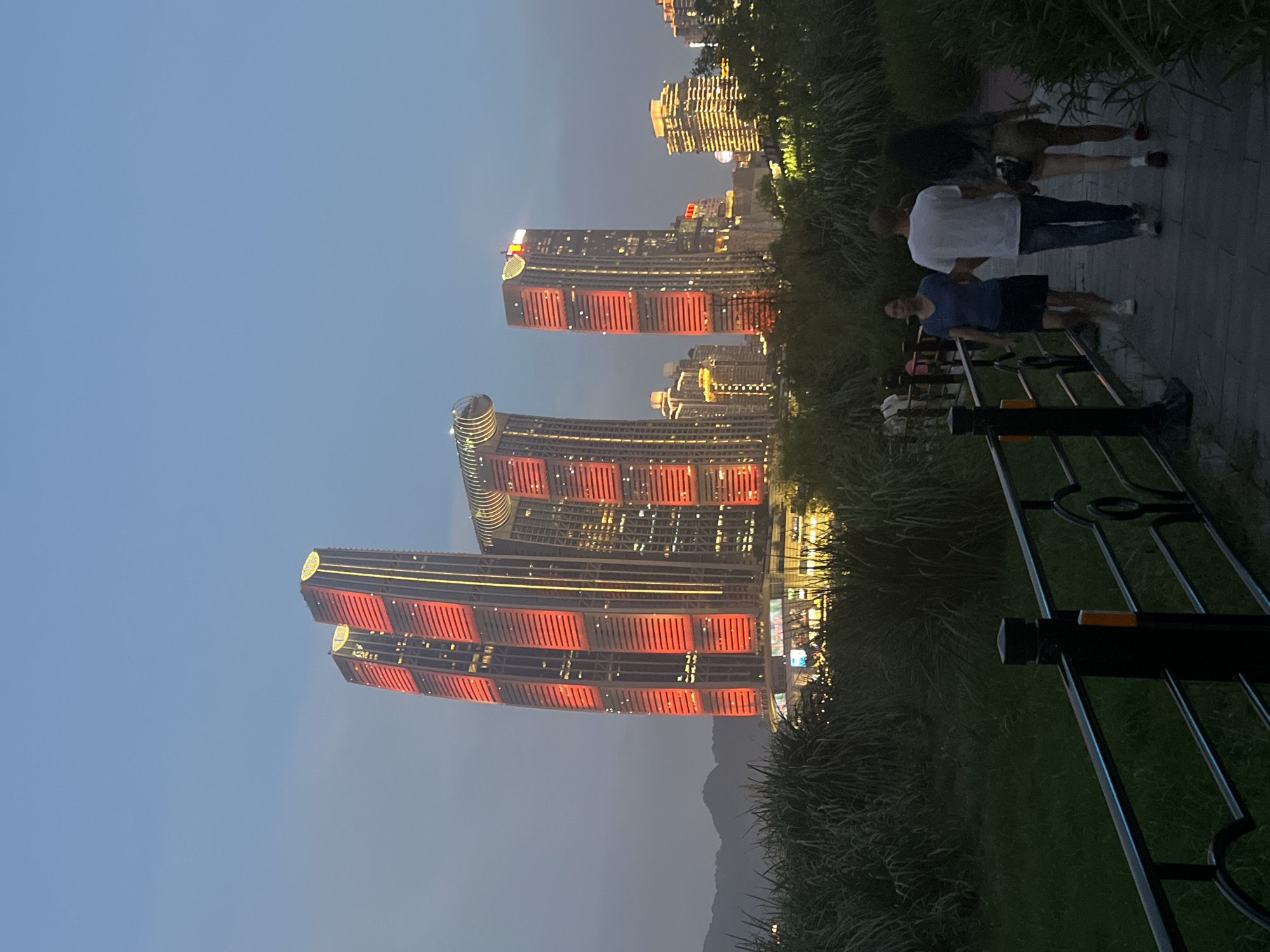 In the twilight, a pathway curves through lush green grass and dense reeds, bordered by a dark metal railing. Three individuals walk along the paved path away from the viewer: a woman in a blue shirt and black skirt, a man in a white t-shirt and blue jeans, and a woman in a gray jacket and tan shorts. In the background, towering skyscrapers are illuminated with vibrant red and yellow lights. The tallest structures feature striking red vertical bands, while others display warm yellow lighting. A distinct building with a curved, glass-enclosed section is visible among the skyscrapers. In the far distance, silhouetted mountains rise against the fading sky. The scene appears to be located in Jiangbeicheng Street, China.