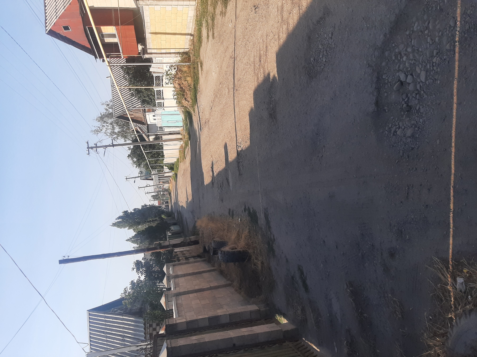 The image depicts a dirt road in the city of Novopokrovka, Kyrgyzstan, lined with residential properties. The road is unpaved and shows signs of wear, with some areas appearing uneven and containing loose gravel. On either side of the road, there are fences and walls made of brick and concrete, separating the properties from the street.
The houses visible are varied in style and material. Some have corrugated metal roofs, while others have tiled roofs. One house in the foreground on the right is painted red and features a balcony. Other houses appear simpler in construction, with some having light-colored walls and white or turquoise gates.
Several utility poles with overhead wires are present along the road, indicating the presence of electricity or communication services. Some of these poles are wooden, while others are metal. Vegetation, including trees and dry grass, is sparsely distributed throughout the scene, particularly along the edges of the road and in front of the properties.
In the foreground on the left, two large tires are visible on the side of the road, partially covered in dry grass. The lighting suggests it is daytime, with clear blue sky overhead. The overall impression is of a rural or semi-urban residential area. There are no people visible in the image.