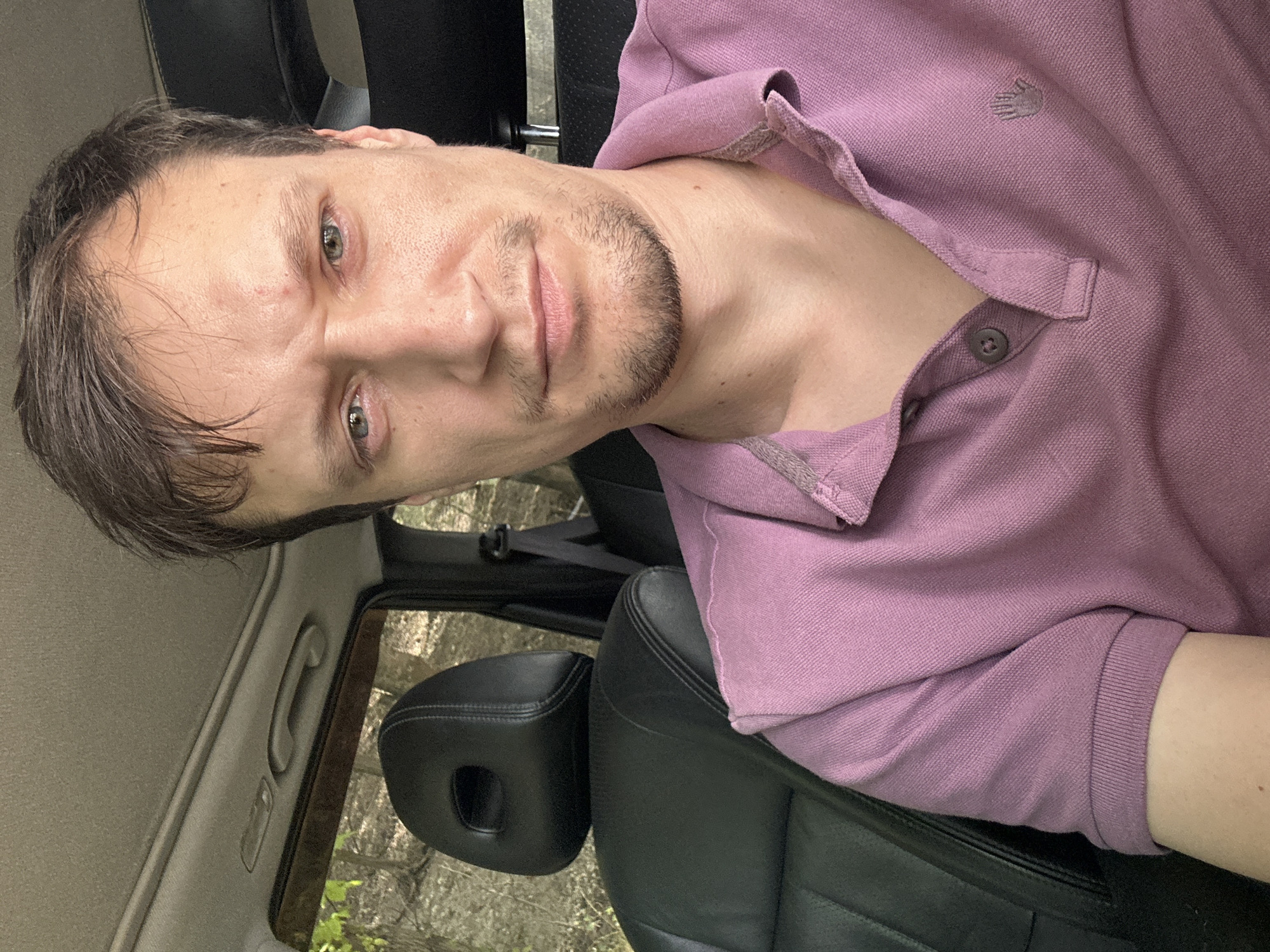 A man with light skin and brown hair is sitting in the driver's seat of a car, looking directly at the camera. He is wearing a pink polo shirt with a collar and two buttons. The interior of the car is visible, including a black leather headrest and seat, a grey headliner, and a grab handle on the ceiling. Through the car window, a wooded area with green foliage is visible. The man appears to have a slight stubble and is making a serious facial expression. The location is identified as Makiv, Ukraine.