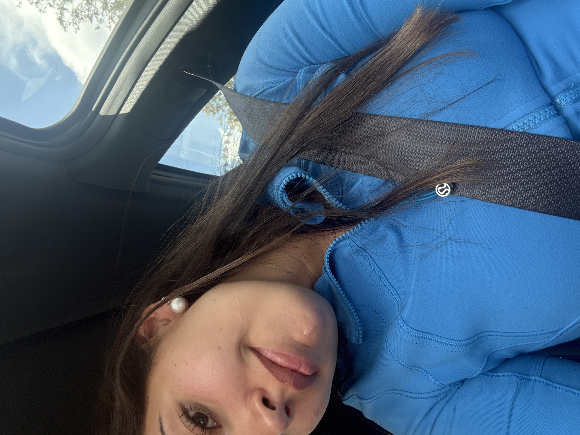 The image depicts a woman wearing a blue jacket and a pearl earring, seated in a car. She is wearing a seatbelt. The car's sunroof shows a view of the sky and tree branches. The image was taken inside a car in Puebla, Mexico.