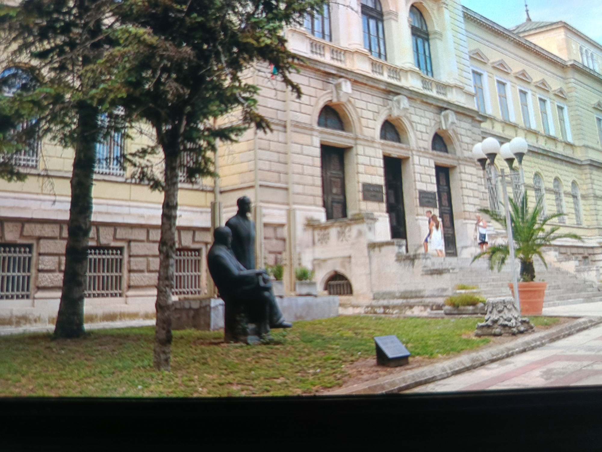 The image captures a view of the historical Varna Courthouse in Varna, Bulgaria. In the foreground, two bronze statues depicting seated figures are prominently displayed on a grassy area with some surrounding trees. One statue is of a man in a suit, seated with his legs crossed and hands resting on his lap. To the right of this statue, another darker, less defined statue is partially visible. A small, dark plaque is situated on the grass to the right of the statues. Behind the statues, the Varna Courthouse building stands, characterized by its ornate architecture, including arched windows, decorative stonework, and a light-colored facade. Several people, appearing as small figures in the distance, are seen near the entrance and on the steps of the building. Three street lamps with multiple globe-shaped lights are positioned in front of the courthouse. A potted palm tree in a large terracotta pot adds a touch of greenery to the scene. The overall impression is that of a public square or park area in front of a significant civic building.