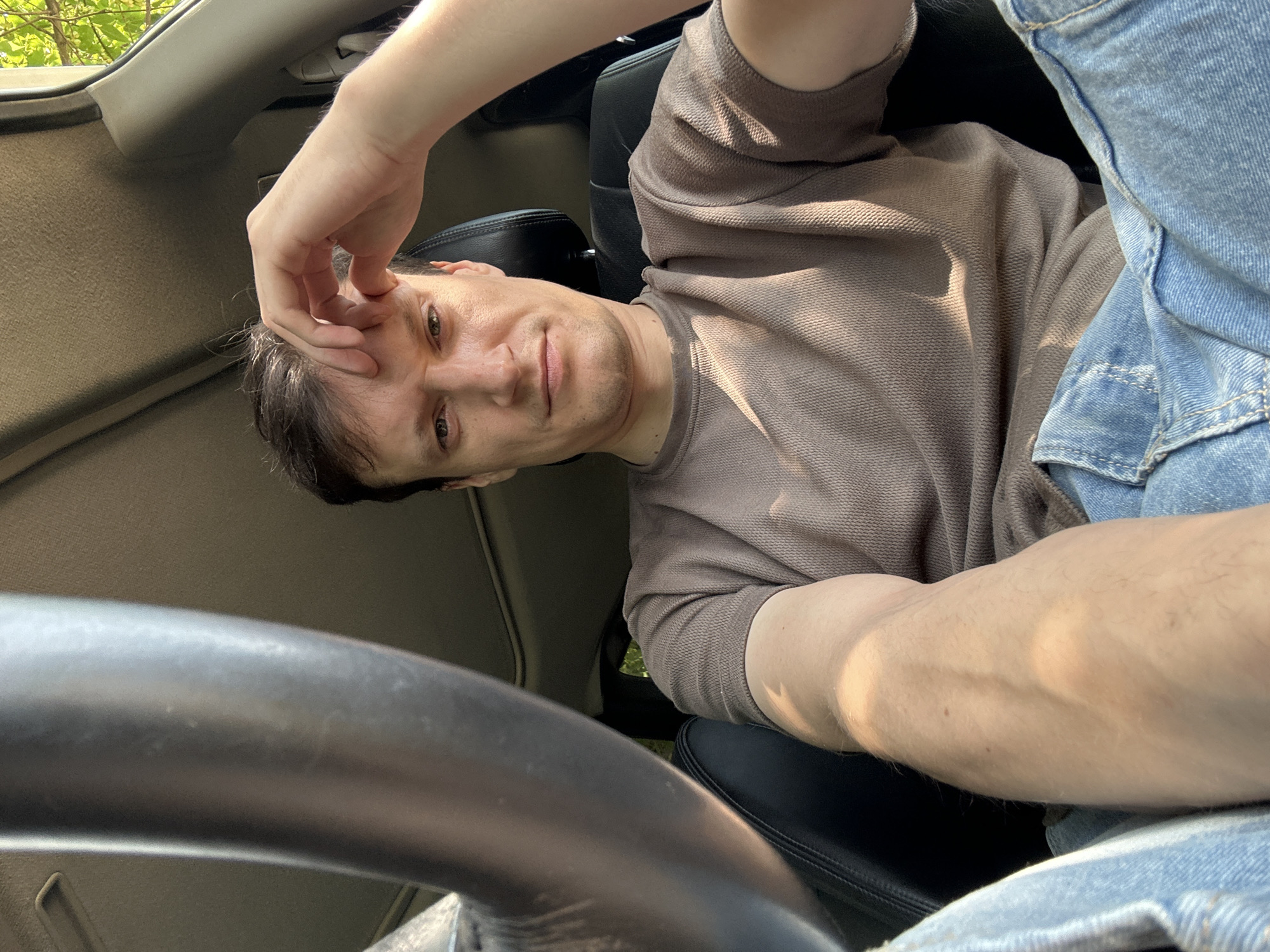 A man is sitting in the driver's seat of a car, with his hand resting on his forehead. He is wearing a brown t-shirt and blue jeans. The car appears to be parked, with sunlight filtering in from the right side, illuminating the interior. The man's expression is difficult to decipher, but he does not appear to be in distress. The interior of the car is partially visible, including the steering wheel and the dashboard. Outside the car, greenery and trees are visible, suggesting an outdoor setting, possibly in a rural or suburban area. The specific location in Makiv, Ukraine, cannot be determined from the image alone.