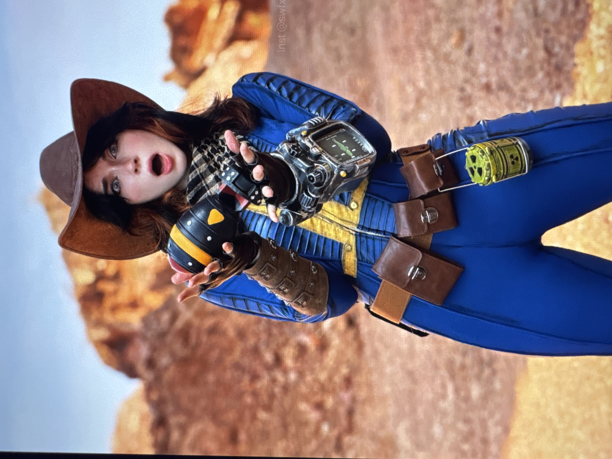 The media file displays a person dressed in a Fallout-themed costume, likely a Vault Dweller. The costume consists of a blue jumpsuit with yellow accents and pleats, a brown cowboy hat, a checkered scarf, brown leather gauntlets and pouches, and a Pip-Boy on the left wrist. The person is holding a spherical object with black and yellow markings, resembling a grenade, in their right hand. A small, cylindrical object with a radiation symbol, also appearing to be part of the costume or a prop, is attached to the belt on the right side.
The person's facial expression is one of surprise or shock, with their mouth open and eyes wide. Their hair is dark and appears to be styled loosely.
The background is out of focus, showing a barren, rocky landscape with a light blue sky, suggesting an outdoor, possibly desert or post-apocalyptic environment. The lighting suggests daytime, and the overall mood conveyed is dramatic due to the person's expression and the costume's elements.
Visible text includes "Inst @swfx" written vertically in white on the background, likely an Instagram handle.