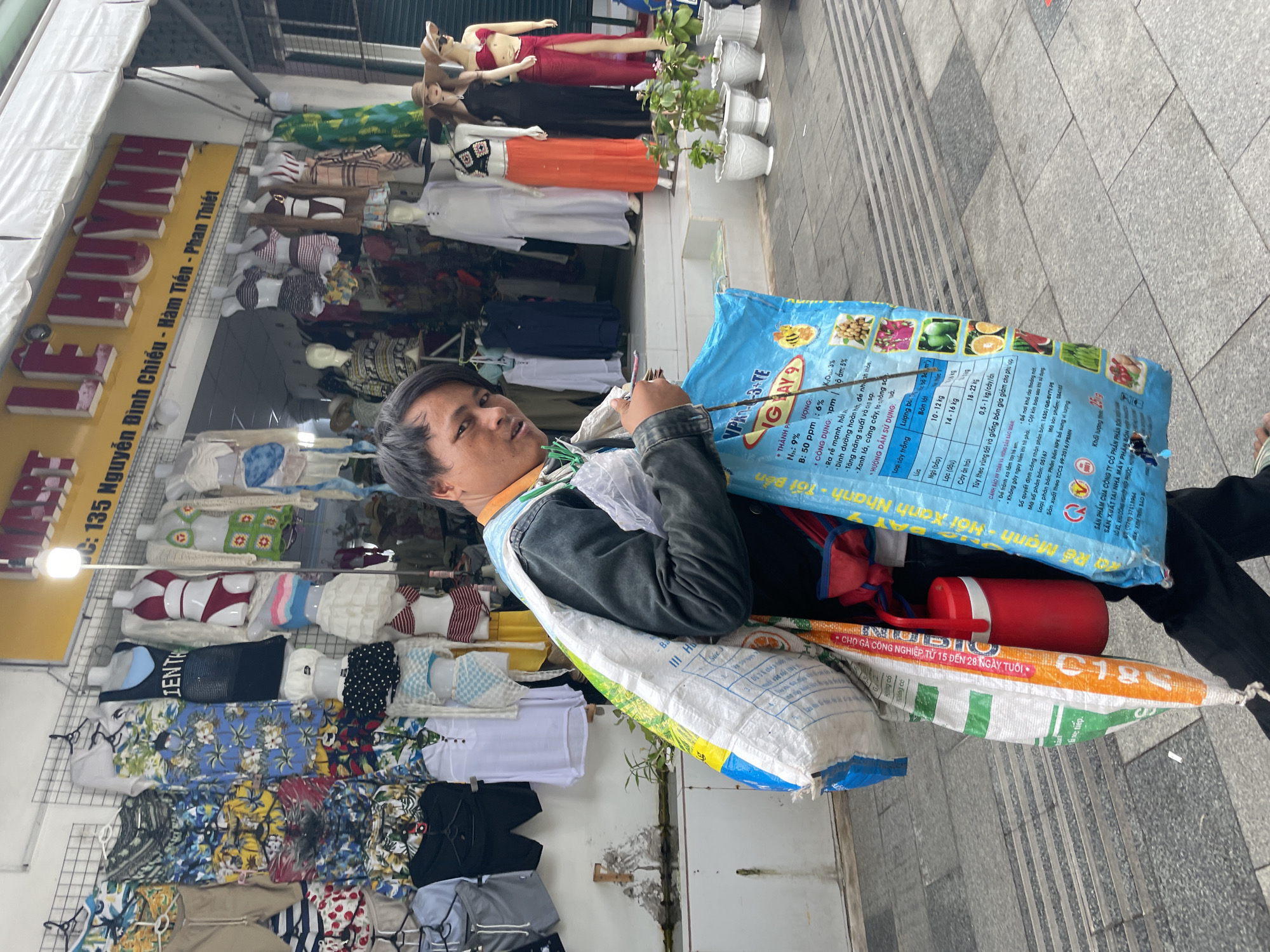 A man stands in front of the "LE HUYNH" clothing store at 135 Nguyen Dinh Chieu, Ham Tien, Phu Thuy Ward, Vietnam, carrying a bag of fertilizer that reads "VPK-3-TE IGA." He also carries a red cylindrical container, a white plastic bag, and a stick. The storefront behind him displays an assortment of clothing on mannequins.