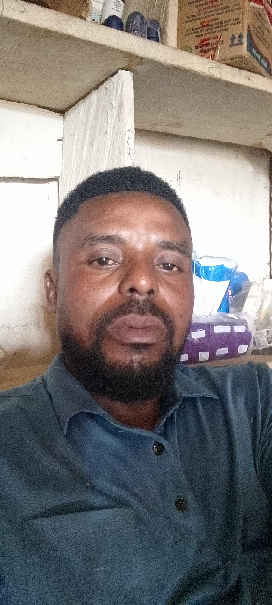 A close-up, waist-up, self-portrait photograph shows a dark-skinned man with short, black hair and a full beard. He is wearing a dark blue collared shirt. The man is looking directly at the camera with a neutral expression.
In the background, there are shelves filled with various items, including boxes and what appear to be plastic bags. The environment seems to be an indoor space, possibly a shop or storage area, with light-colored walls and shelving units. The lighting is natural and appears to be coming from the front, illuminating the man's face.
The image was taken in Durumi, Nigeria.