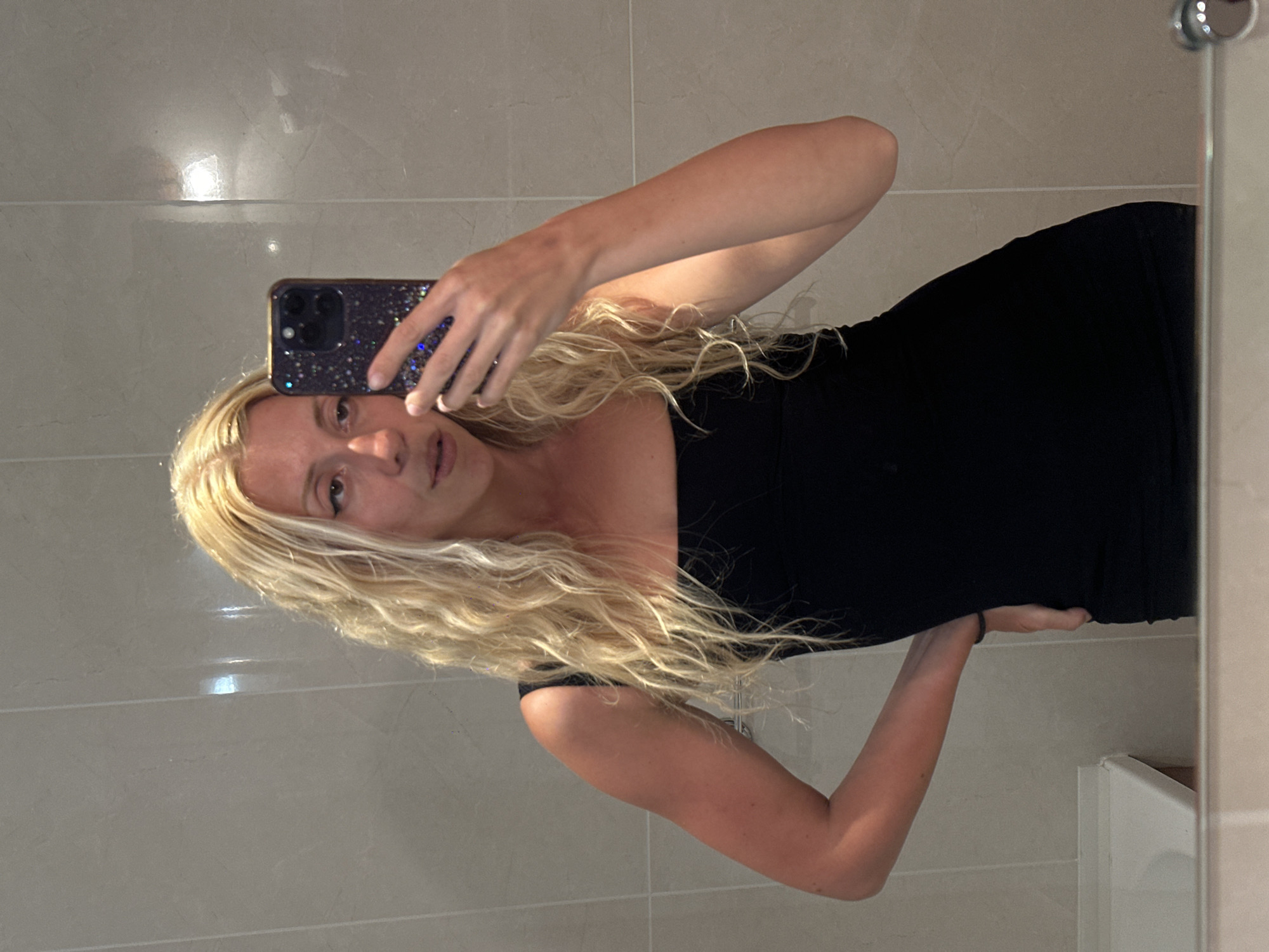 A blonde woman in a black dress takes a selfie in a bathroom mirror. The bathroom has light beige tiled walls. The woman is holding a dark smartphone with a sparkly case. Her left hand is on her hip. The bottom of a white sink is visible in the lower left corner of the image, reflected in the mirror. The scene is indoors, likely in a domestic or hotel setting. The location of Polincove, France is not evident from the image.