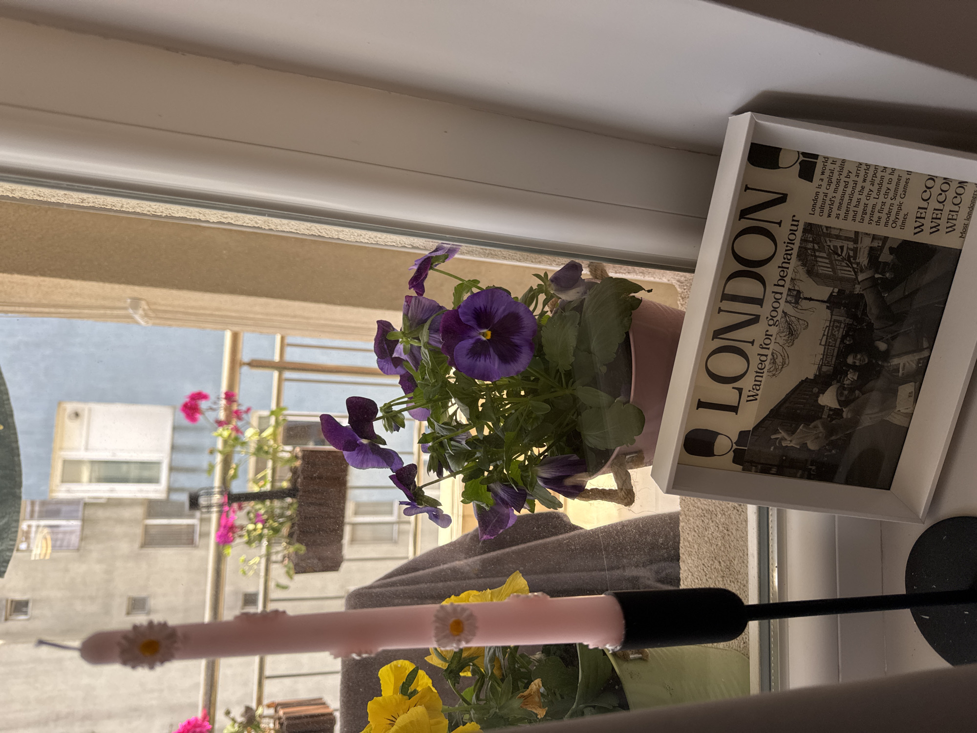 The image depicts a windowsill decorated with plants and a framed print. A pot of purple pansies sits prominently, surrounded by other plants with yellow and pink flowers, reflecting the view outside. The framed print, angled towards the viewer, displays the word "LONDON" in bold capital letters above a black and white image. The print's text describes London as a world cultural capital. A decorative pink object with daisy embellishments rests in the foreground. The windowsill is located in Burgas, Bulgaria.