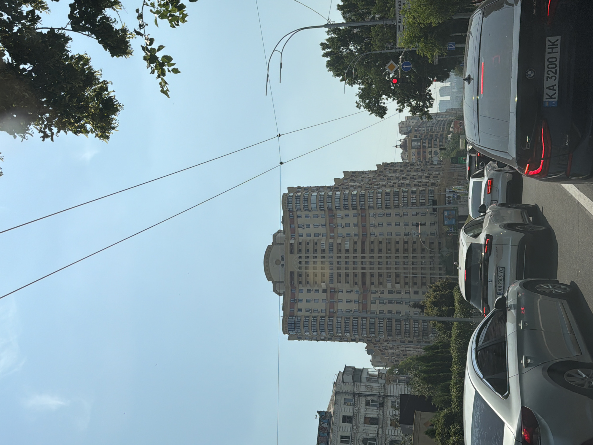 The image shows a street scene in Kyiv, Ukraine, likely on a sunny day. Several cars are stopped in traffic, including a silver sedan in the foreground on the left, a white SUV behind it with license plate AA 2306 YC, and a black BMW sedan with license plate KA 3200 HK in the foreground on the right. In the background, there are several tall, multi-story buildings, including a distinctive curved building with many windows. Trees line the street, and overhead power lines are visible. Traffic lights and street signs are also present. The sky is clear and blue.