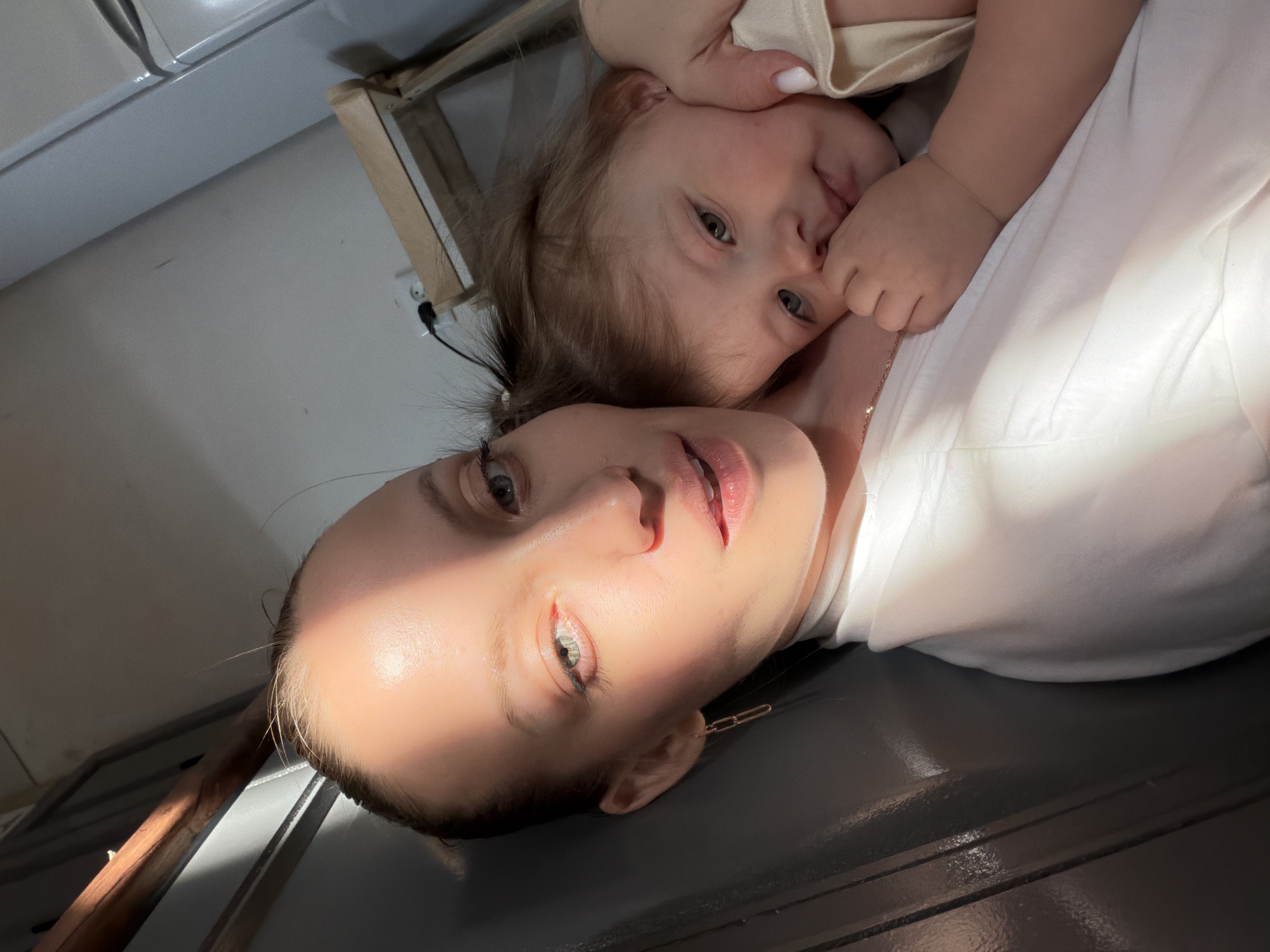 An adult female, appearing in the foreground with light-colored hair pulled back, holds an infant in an indoor setting in Reshetnikovo, Russia. The female wears a white high-necked top, a thin gold chain necklace, and a gold link earring in her left ear. Her face, partially illuminated by a strong diagonal beam of natural light, displays blue-green eyes with light blue eyeliner and is directed towards the camera. The infant, with light-colored hair and blue-green eyes, rests its head on the female's right shoulder, wearing a light-colored top. One of the infant's hands is near its mouth/chin, and its gaze is directed slightly downwards and to the right. The adult female's left hand, featuring white nail polish, supports the infant's head area. The background on the left consists of a dark, glossy, possibly panelled surface. The background on the right includes a light-colored wall with a black electrical cord plugged into a white wall socket, a light wooden furniture frame, and a portion of a white vertical appliance.