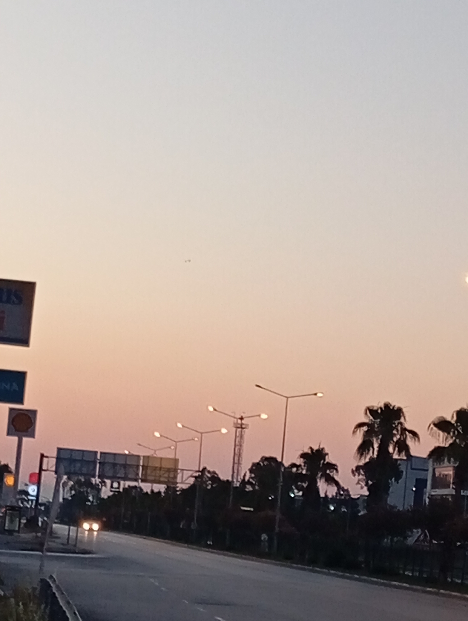 This media file captures a street scene at dusk in Konyaaltı, Turkey. The sky is a gradient of pale yellow and pink, indicative of sunset. In the foreground, a road curves gently to the right. Two sets of headlights from an oncoming car are visible in the distance, their bright beams cutting through the fading light.
Along the roadside, several streetlights are illuminated, casting a soft glow. Palm trees and other dark foliage line the background, silhouetted against the colorful sky. Behind the trees, buildings are partially visible, including a structure that appears to be a factory or industrial complex. A traffic light shows a red signal on the left side of the road.
There are several signs visible. One prominent sign on the left has the word "US" in large letters on a dark background. Below it, another sign has the word "NA" in white letters on a blue background. A circular sign with a yellow and red logo, possibly representing a fuel station, is also present. In the distance, a tall lattice tower, likely for communication or power, can be seen. The overall mood is peaceful, with the gentle light of sunset and the quiet hum of a road at the end of the day.