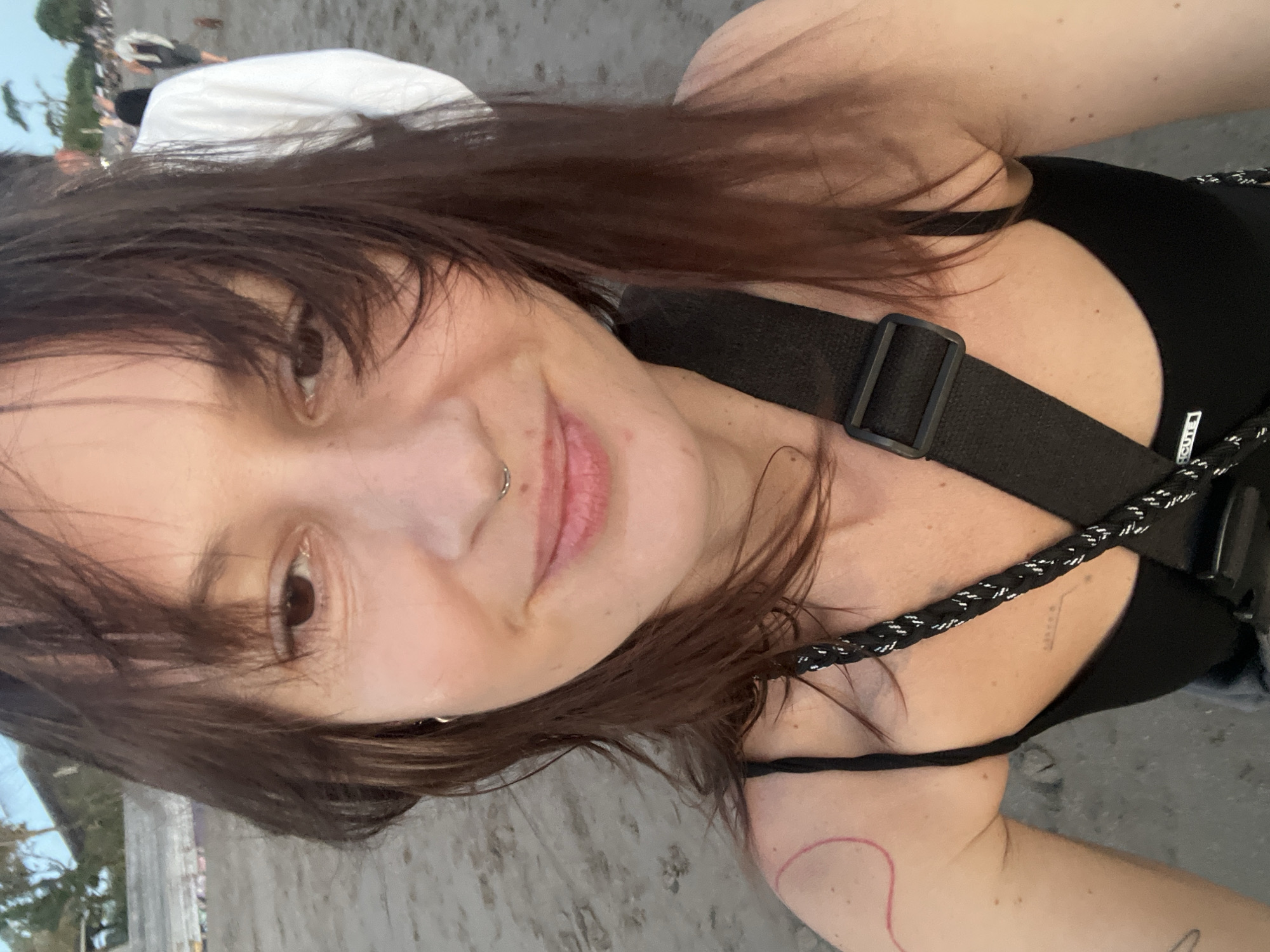 A selfie captured in North Kuta, Indonesia, shows a fair-skinned woman with brown hair and a nose piercing. She is wearing a black top with straps. A black strap, possibly from a bag or harness, crosses her body. A faint red circle is visible on her arm. In the background, indistinct figures and a white object suggest an outdoor, possibly public, setting. The ground appears to be sand.