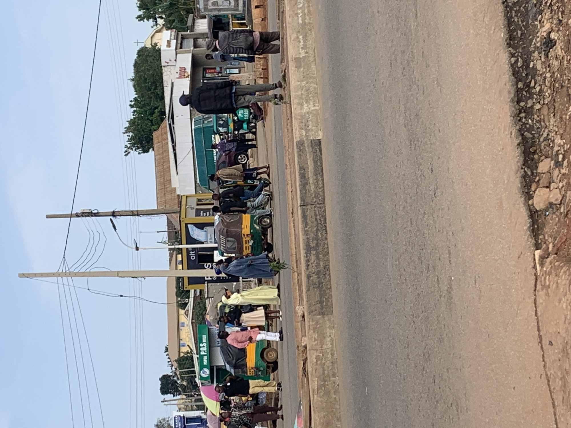 The image depicts a street scene in Gura Top, Nigeria, likely a busy intersection or market area. Several people are visible, some walking, others standing in groups. A prominent feature is a row of small shops or stalls, one of which has a "POS Outlet" sign. Several auto-rickshaws, commonly known as "keke napep" in Nigeria, are present, indicating public transportation services. A green minivan is also parked on the street. The environment appears to be a typical urban setting with overhead electrical wires and poles. The road surface shows signs of wear and tear. The overall impression is one of daily life and commerce in a Nigerian city.