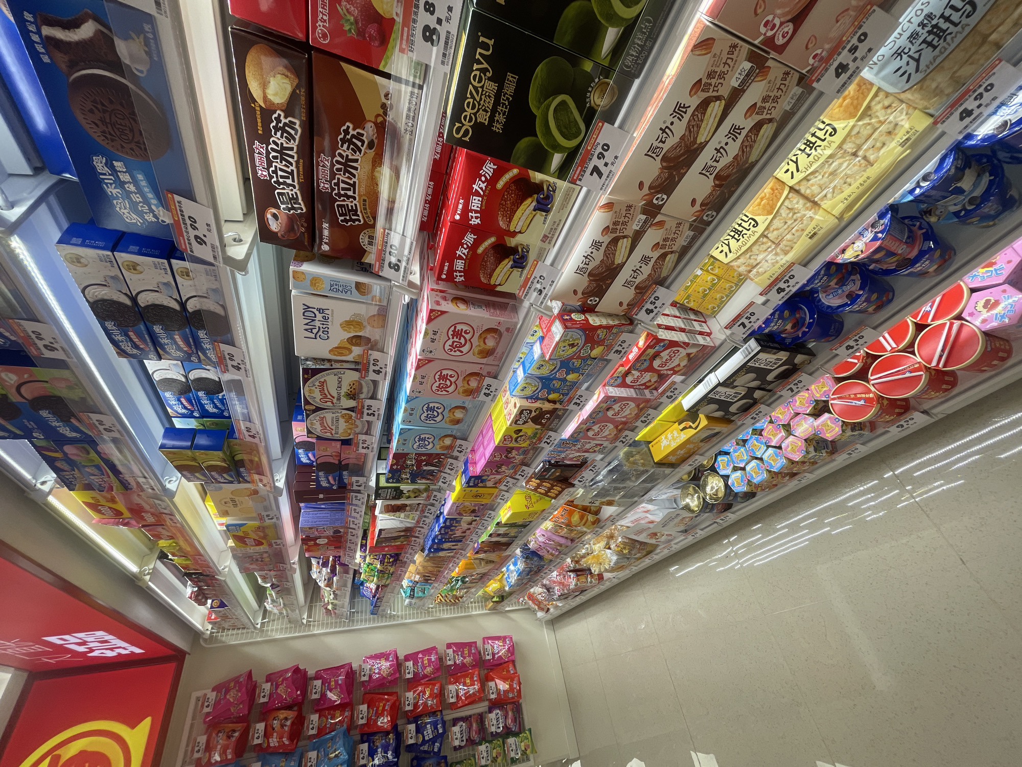 The image shows a brightly lit, multi-tiered shelf display in what appears to be a convenience store or supermarket. The shelves are filled with a variety of packaged snacks, predominantly cookies, biscuits, and cakes. Many of the products have vibrant packaging with Chinese text. Prominently displayed are boxes of "OREO" cookies, "LANDY CASTLE" cookies, "提拉米苏" (Tiramisu) flavored cakes, and "Seezyu" matcha-flavored cakes. Other items include small bags of snacks, cans of what seem to be cookies or crackers, and brightly colored snack tins at the bottom of the display. The overall impression is of a well-stocked snack aisle. The floor is tiled with a light-colored material, reflecting the overhead lighting. No people are visible in the frame.