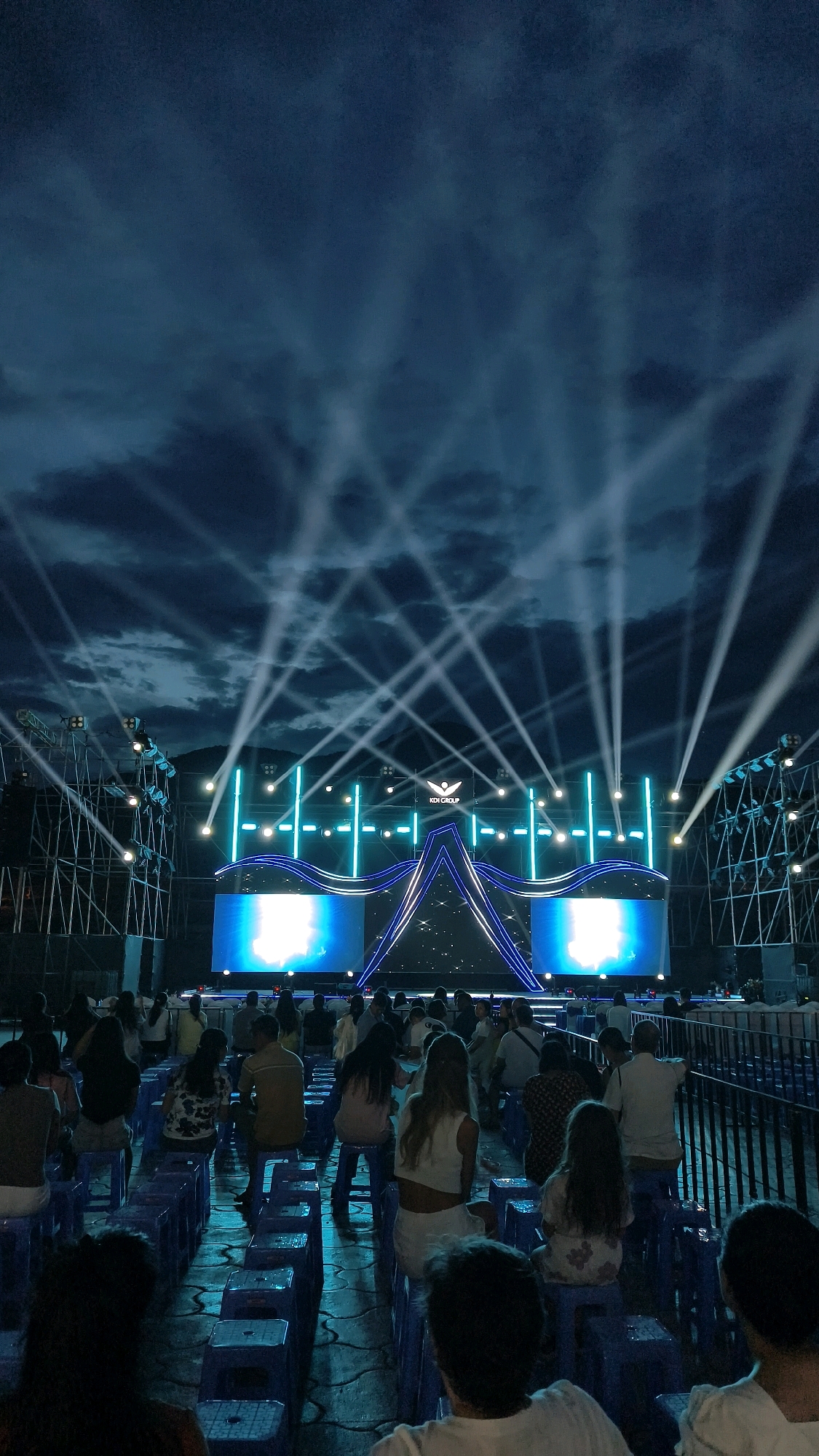 An outdoor stage setup in Nha Trang, Vietnam, is illuminated by numerous spotlights against a cloudy twilight sky. A crowd of people sits on blue plastic stools facing the stage, which features large LED screens and elaborate blue and white lighting designs. The stage displays the logo "KDI GROUP". The overall atmosphere suggests a nighttime event or concert.