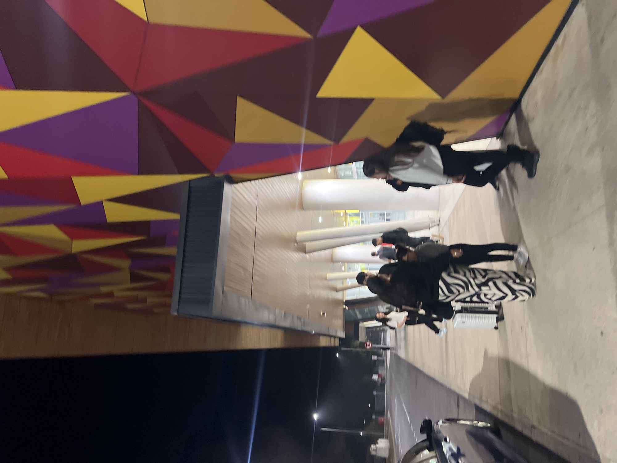 The image depicts a group of people with luggage outside a building with a brightly colored geometric facade. The wall features triangles in shades of yellow, purple, and red. Several people are walking away from the building. A car is visible on the street, parked to the right of the group. Streetlights illuminate the scene. The location is Arica, Chile.