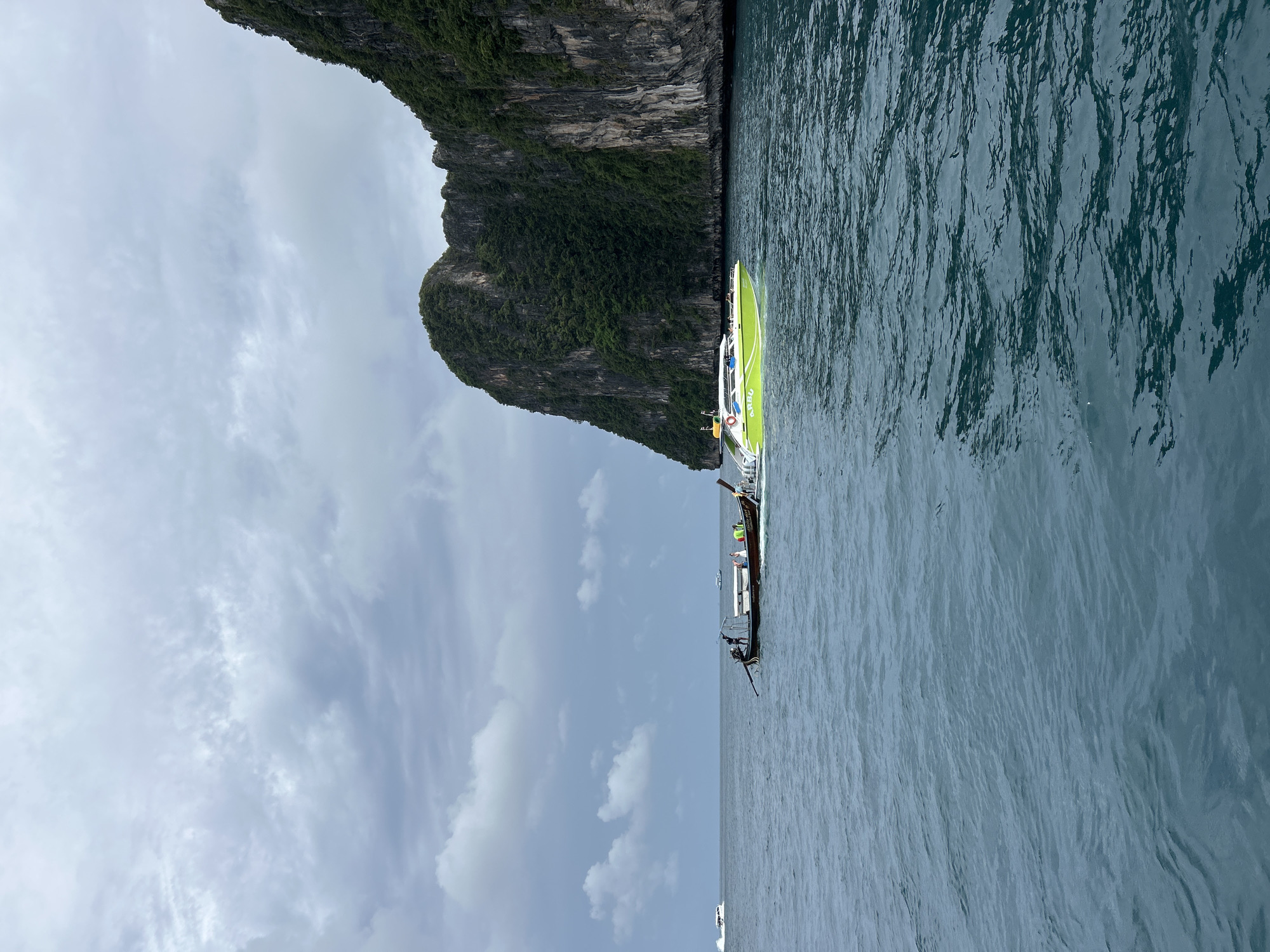 The image depicts a scenic view of the waters near Ao Nang, Thailand. In the foreground, the deep blue sea is calm with small ripples. Two boats are visible on the water. A traditional longtail boat, with a wooden hull and a characteristic long propeller shaft, is positioned towards the left of the frame. Several people are seated inside this boat. To its right, a brighter, modern speed boat with a lime green hull and white canopy is moving. The speed boat has the word "ARBU" visible on its side and a life preserver is attached to its railing.
In the background, a large, towering limestone cliff covered in lush green vegetation rises dramatically from the sea. The cliff face is steep and rocky. The sky above is overcast with a mix of grey and white clouds, creating a diffused light. In the far distance, on the left side of the image, a white boat is also visible. The overall scene captures a typical day of marine activity and the striking natural beauty of the Ao Nang coastline.
