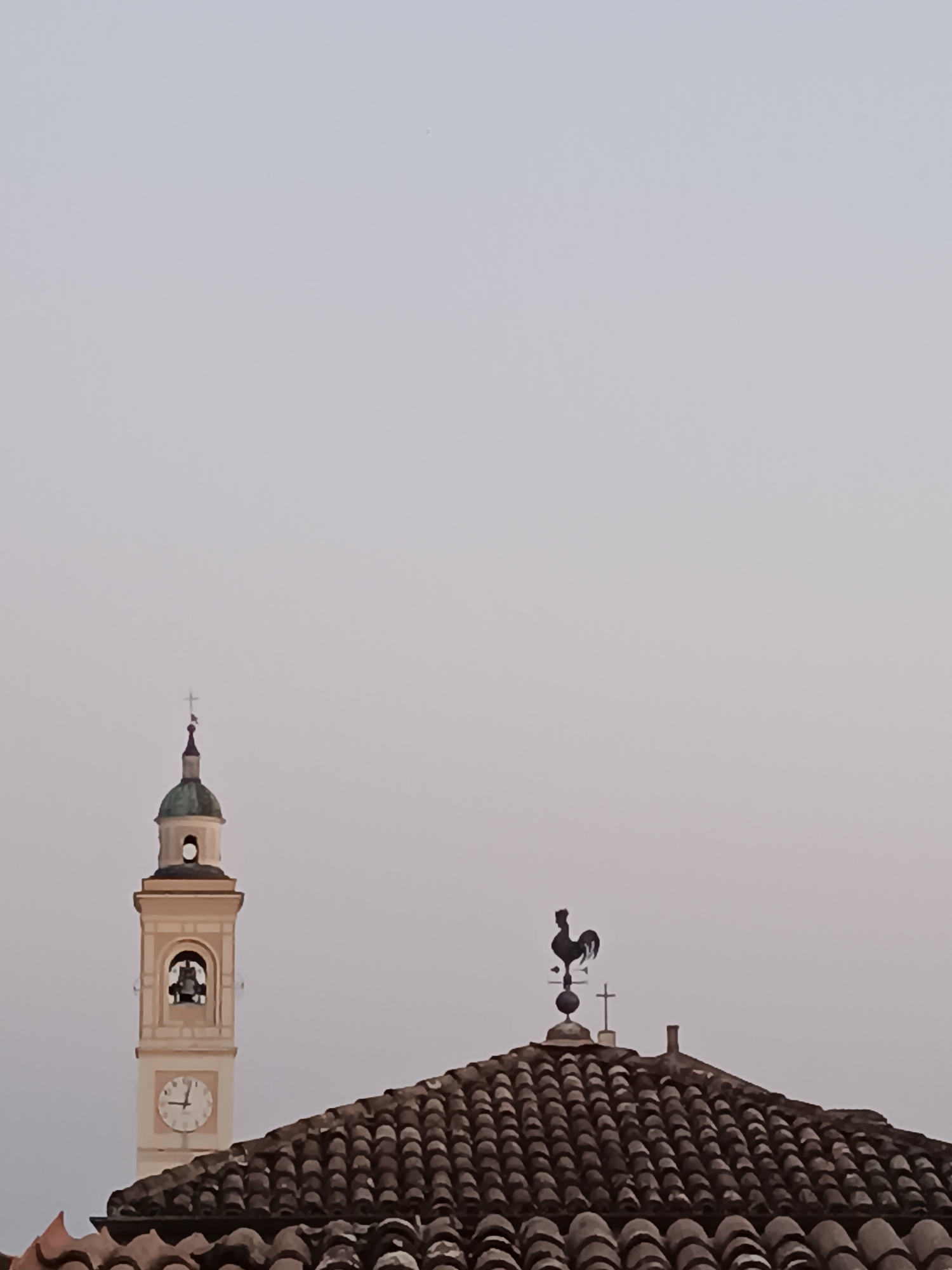 The image captures a view of a bell tower and a tiled roof against a pale sky. The bell tower, light-colored and with a green dome, features a bell visible through an opening and a clock face showing approximately 1:40. To the right, a rooster-shaped weather vane stands on a separate tiled roof. The scene is likely from Viadana, Italy, given the context.