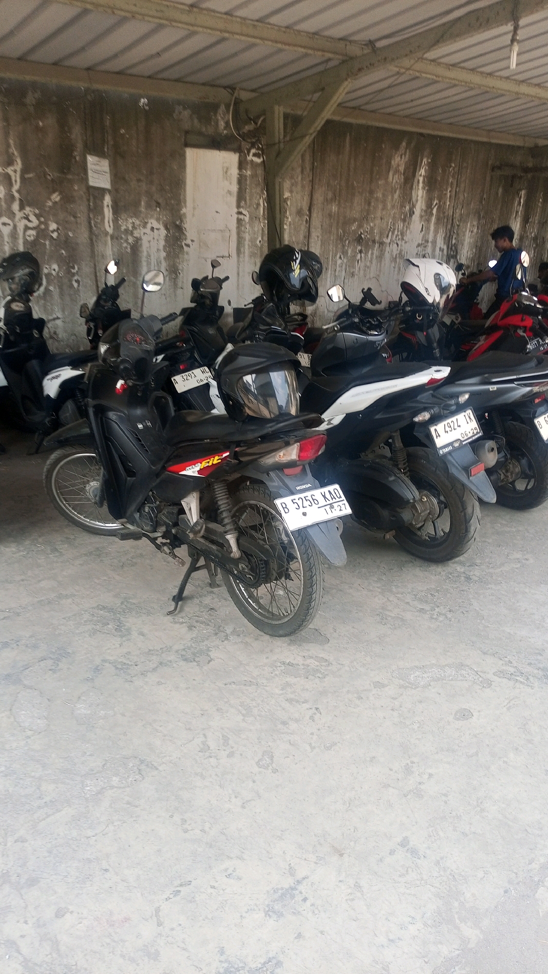 An outdoor photograph depicts a line of parked motorcycles under a covered, weathered concrete structure with a corrugated metal roof. The motorcycles are primarily black and red. In the foreground, a black motorcycle with a license plate reading "B 5256 KAO" is parked on a textured concrete ground. Several other motorcycles are visible behind it, some with helmets placed on them. To the right, a person wearing a blue shirt is interacting with one of the motorcycles. The background consists of a textured, weathered concrete wall. The overall scene suggests a parking area for motorcycles. The image is a landscape orientation with a slightly overcast sky visible in the background.