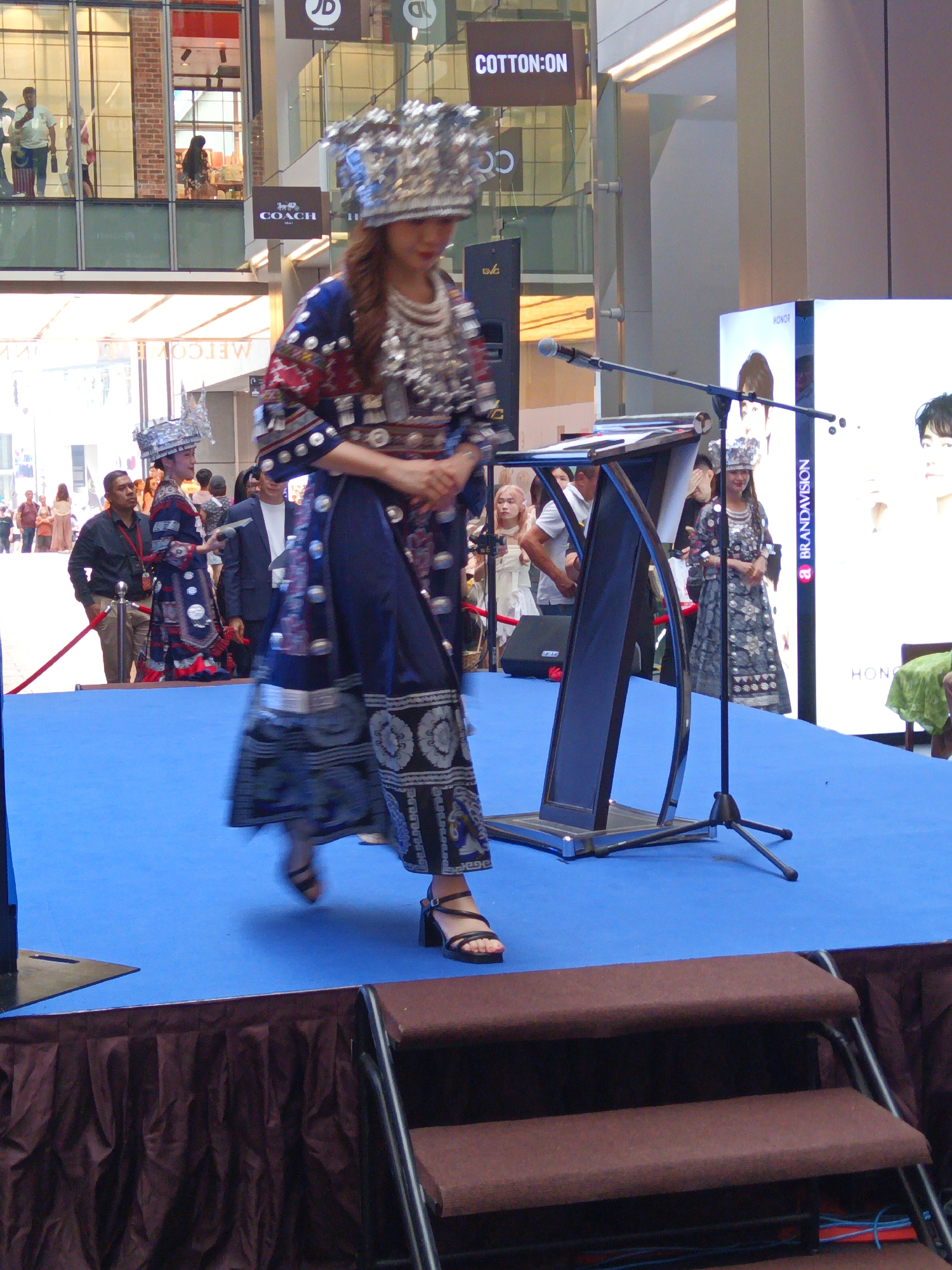 A woman in traditional ornate clothing and headwear is on a blue stage, likely performing or presenting. She is wearing a silver, intricate headdress and a dark blue and patterned dress with red accents. Her arms are crossed in front of her, and she is looking down. To her left, another woman in similar traditional attire stands facing away from the camera. In the background, there are several people, including men in suits and women in casual clothing. A podium with a microphone stands on the stage. Behind the stage, a large digital screen displays portraits of individuals. The scene appears to be set in a public indoor space, possibly a shopping mall or convention center, with storefronts visible through large glass windows in the background. The location is identified as Pavilion Kuala Lumpur, Malaysia, based on contextual clues such as the "COTTON:ON" signage and the overall mall architecture.
