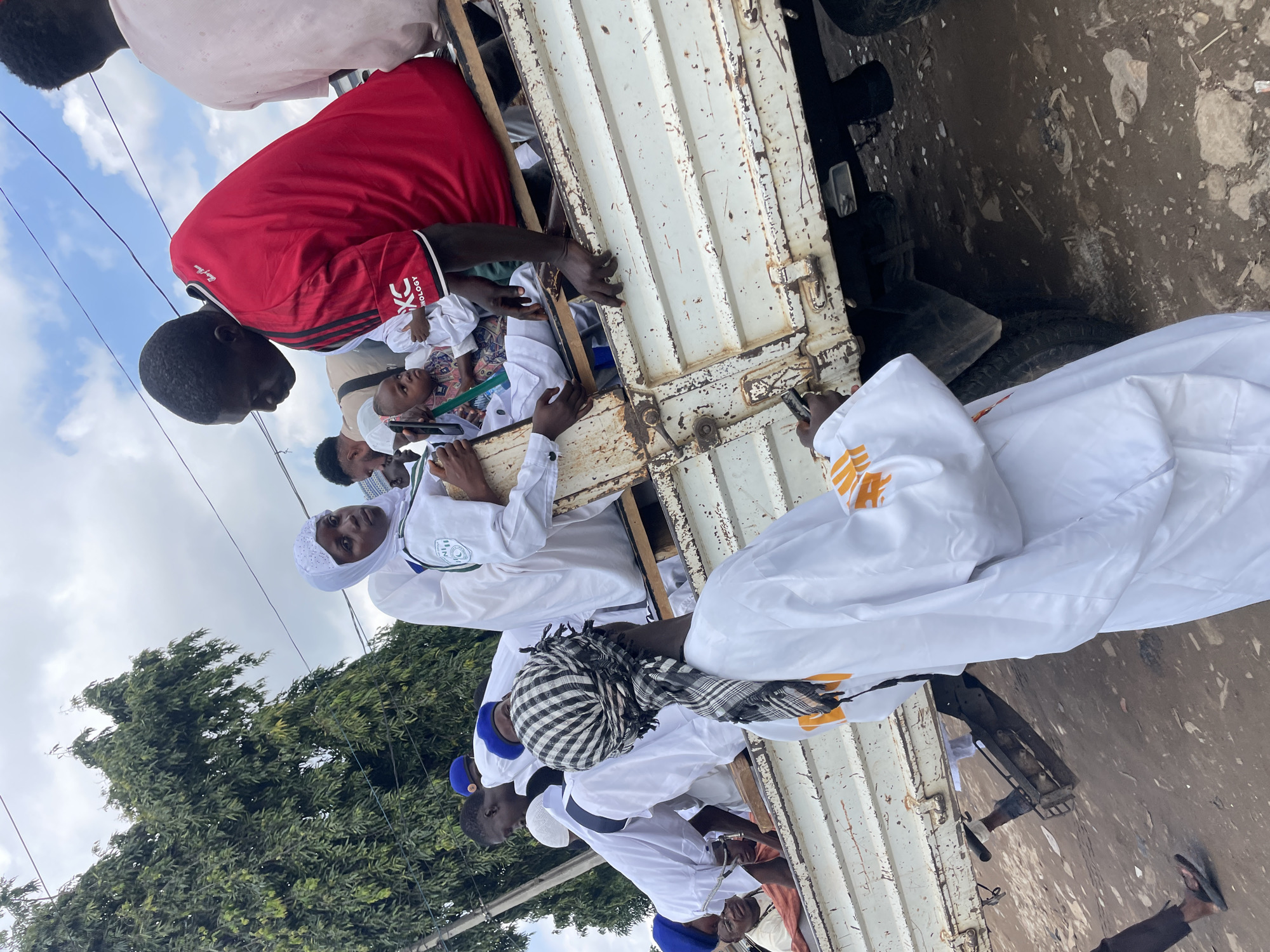 A group of people, most of whom are wearing white garments and head coverings, are gathered around and on the back of a white flatbed truck. The truck appears to be parked on a dirt or unpaved road, with debris scattered on the ground. In the background, there are trees and a cloudy sky. The overall scene suggests a communal gathering or event.