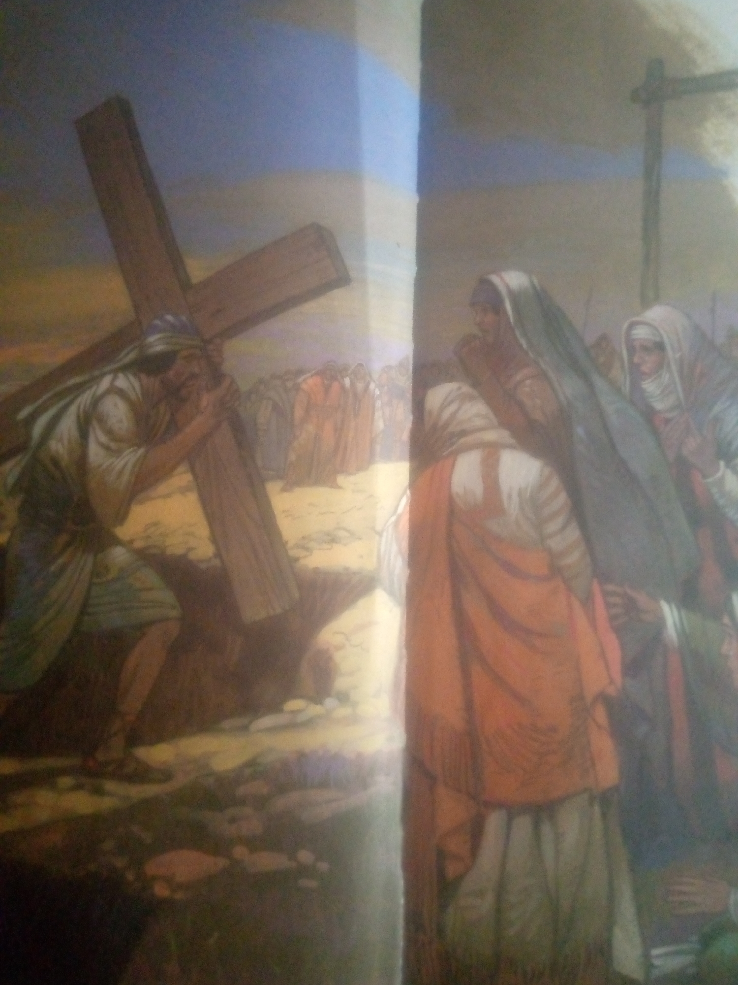 This is a painting depicting a religious scene, likely the carrying of the cross. On the left, a man with a blue head covering is straining to carry a large wooden cross. He is wearing a white tunic and a blue sash. Behind him, a crowd of people in tunics are visible. On the right, several figures are draped in robes, with two women in the foreground wearing head coverings. One woman is in an orange robe, and the other wears a white and blue garment. The background shows a cloudy sky and a barren landscape. The image does not provide information about the city of Jauro Yinu, Nigeria.