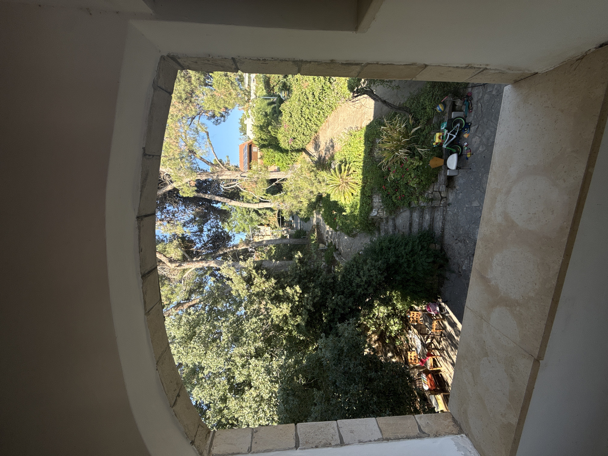 This is a photograph taken from an arched window opening onto a sun-drenched garden in Kfar Vradim, Israel. The foreground is dominated by a light-colored stone windowsill. Beyond this, a lush garden unfolds, characterized by mature trees, dense shrubbery, and flowering plants. A paved pathway winds through the garden, interspersed with stone steps. In the lower left of the garden, a wooden table with chairs is set up, suggesting an outdoor seating area. Further into the garden, a collection of children's toys is visible, including a bicycle, a scooter, and various colorful items scattered on the ground. A partially visible building with a reddish-brown roof is nestled amongst the trees in the background. The sky is clear and blue.